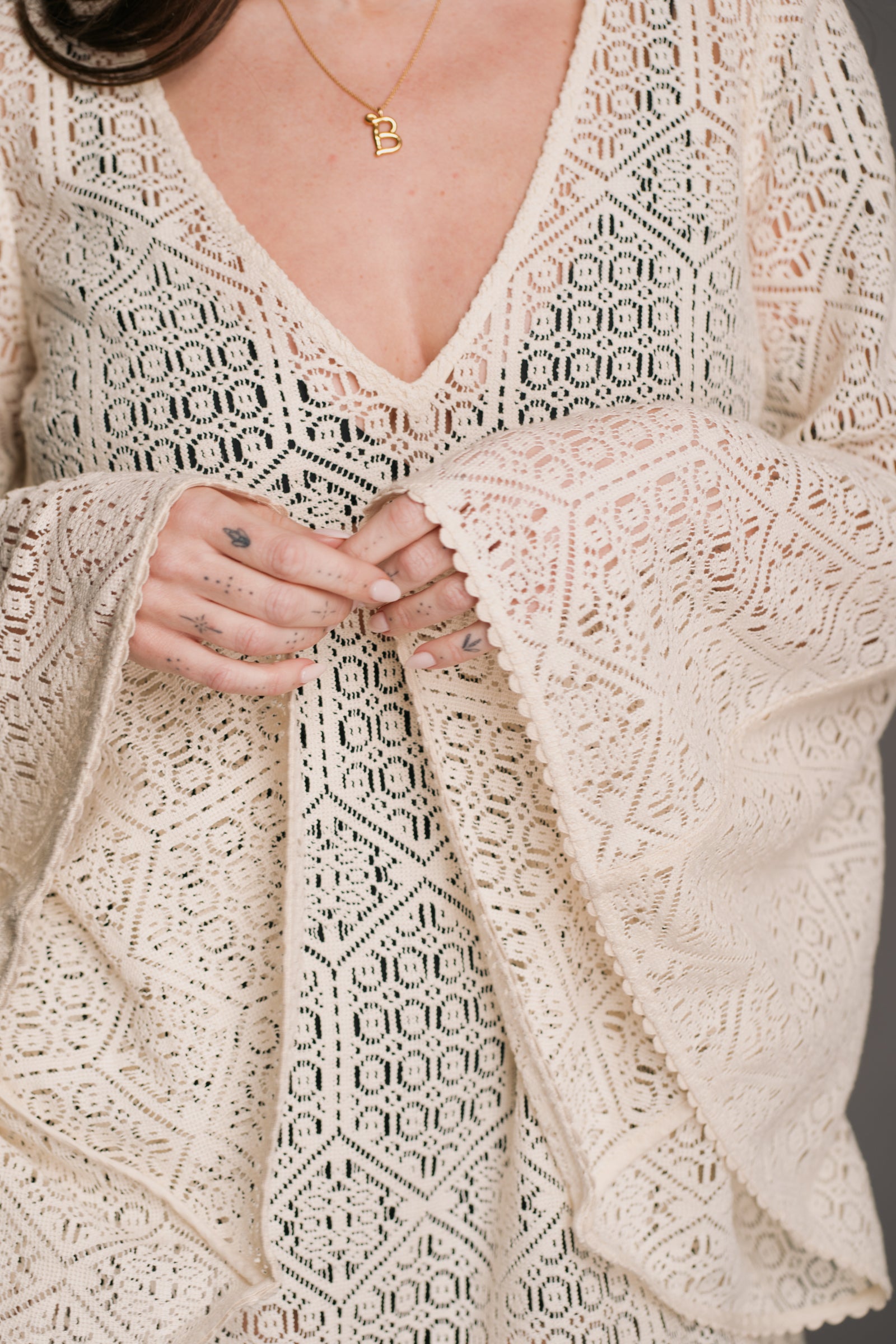 Our model, Bex, standing against a neutral backdrop in the studio wearing the Little Lies Goldie Ivory Crochet Bell Sleeved Mini Dress, featuring intricate crochet detailing, flowing bell sleeves, a V-neckline, and a flattering mini silhouette. Styled with gold 'B' necklace and featuring her intricate hand tattoos - the look embodies effortless bohemian elegance.
