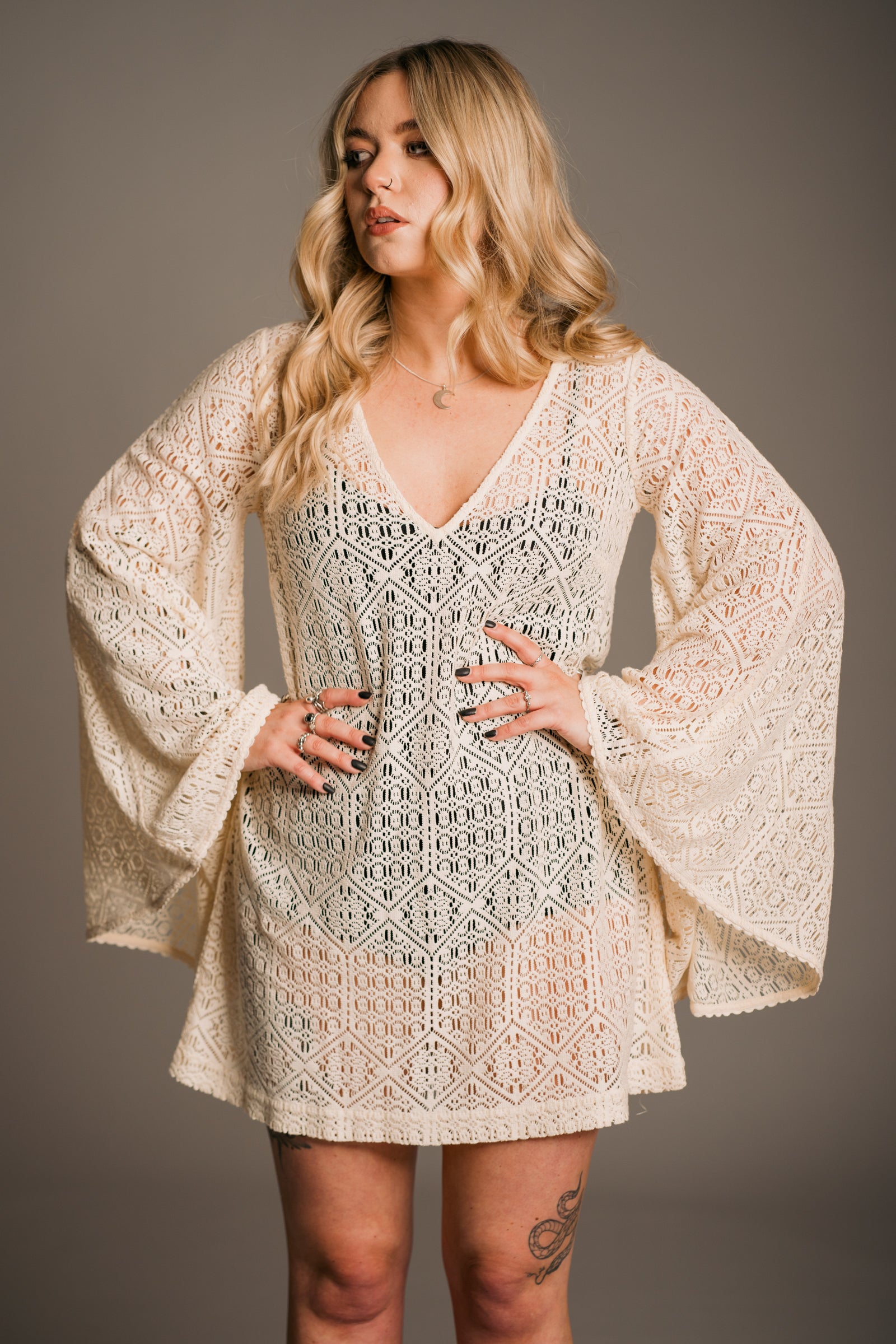 Our model, Erin, standing against a neutral backdrop in the studio rocking wavy blonde hair and wearing the Little Lies Goldie Ivory Crochet Bell Sleeved Mini Dress, featuring intricate crochet detailing, flowing bell sleeves, a V-neckline and a flattering mini silhouette. Styled with Little Lies Sterling Sliver crescent moon necklace and visible leg tattoos - effortless bohemian elegance.