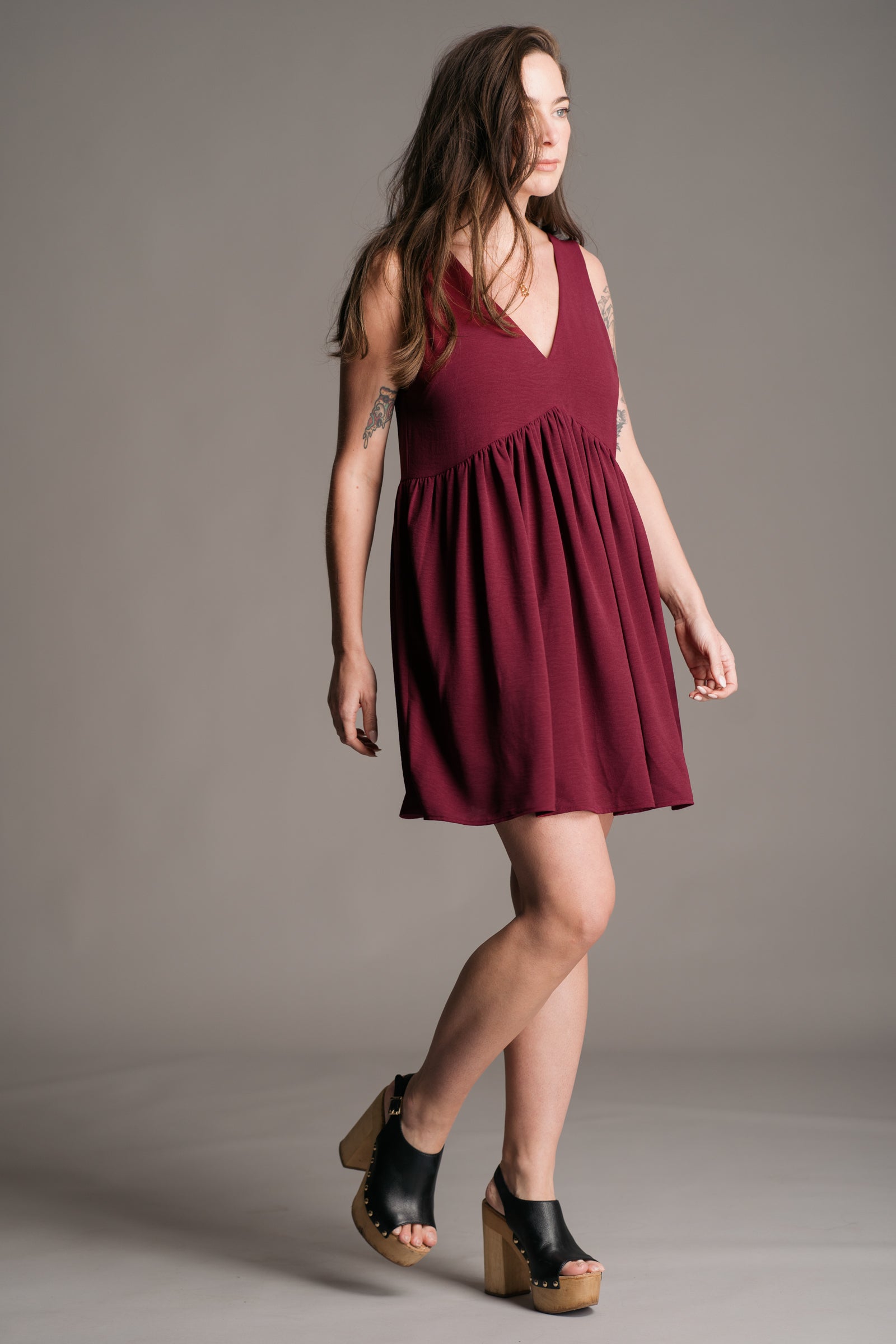 Our model, Bex, wearing the Little Lies Grace Plum Smock Dress, a floaty plum-coloured smock dress with a relaxed fit, styled with black wooden platform clogs. Posing against a neutral backdrop, the outfit exudes effortless bohemian charm.