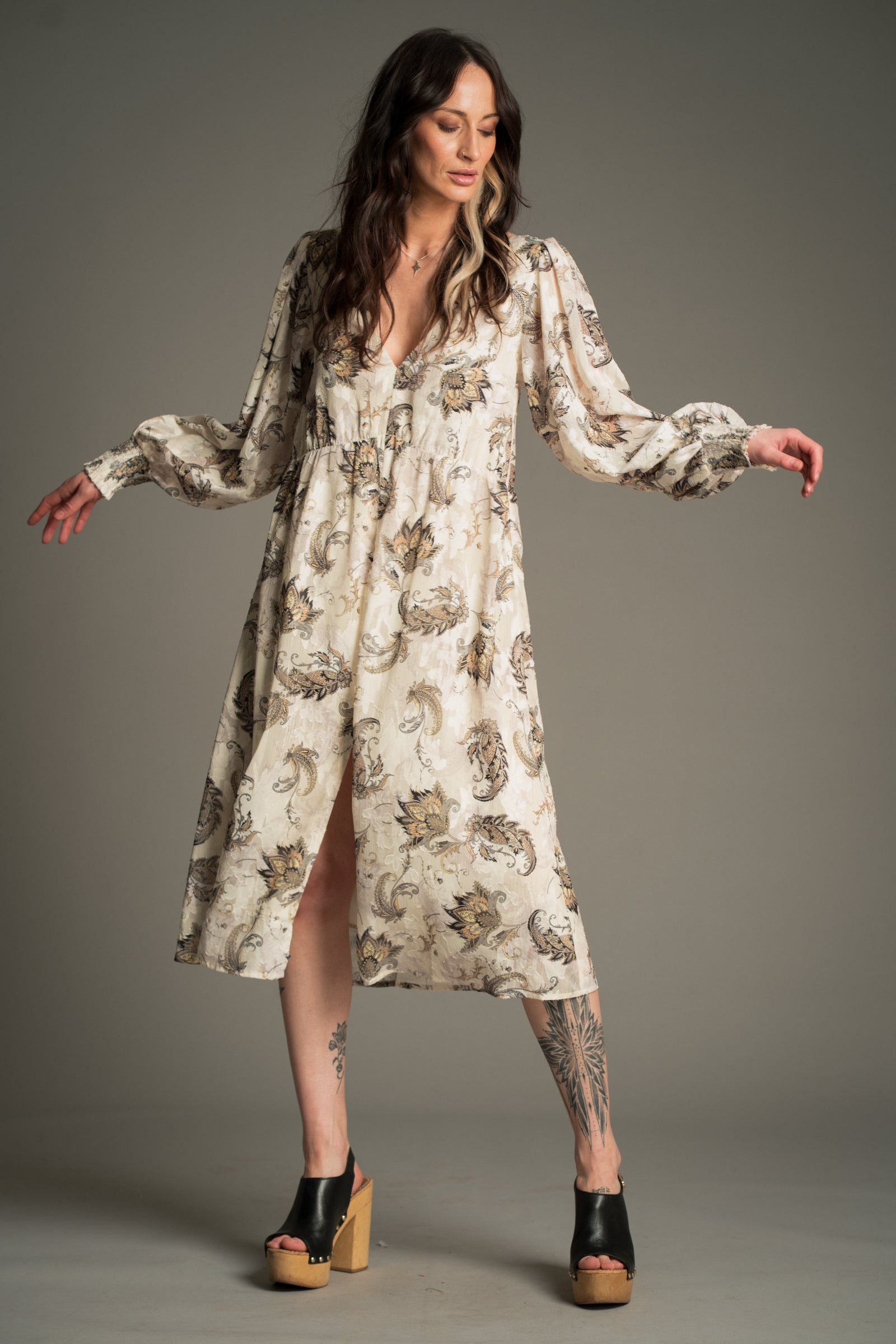 Victoria wears Little Lies Harvest Moon Paisley Midi Dress in cream with an intricate paisley print, full‑length front view featuring long voluminous sleeves, a deep V‑neckline, and a front split hem, styled with platform sandals.