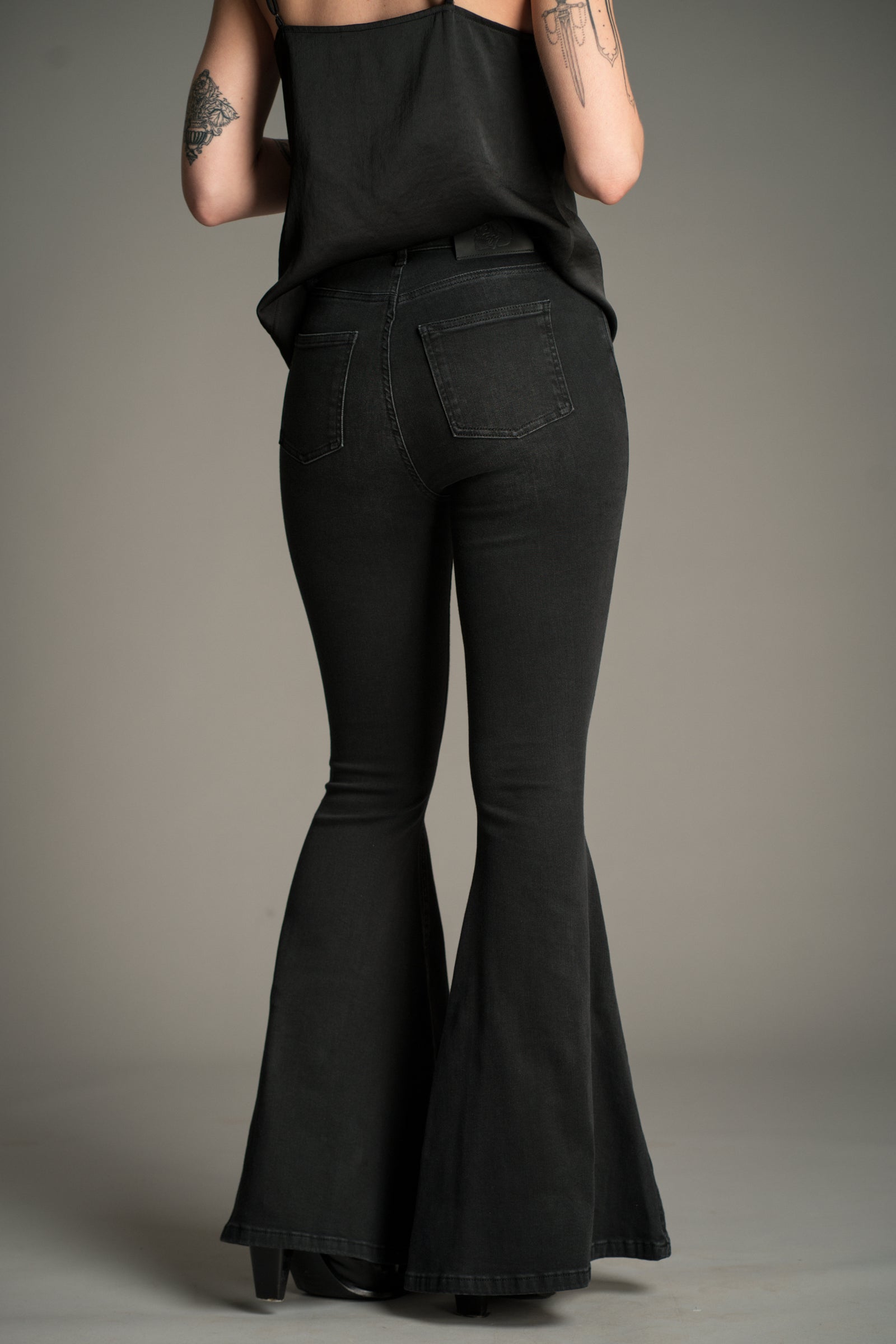 Little Lies Hendrix Denim Flares in washed black denim, back view showing high‑waist fit, rear patch pockets, and wide flared legs, styled with a black satin cami.