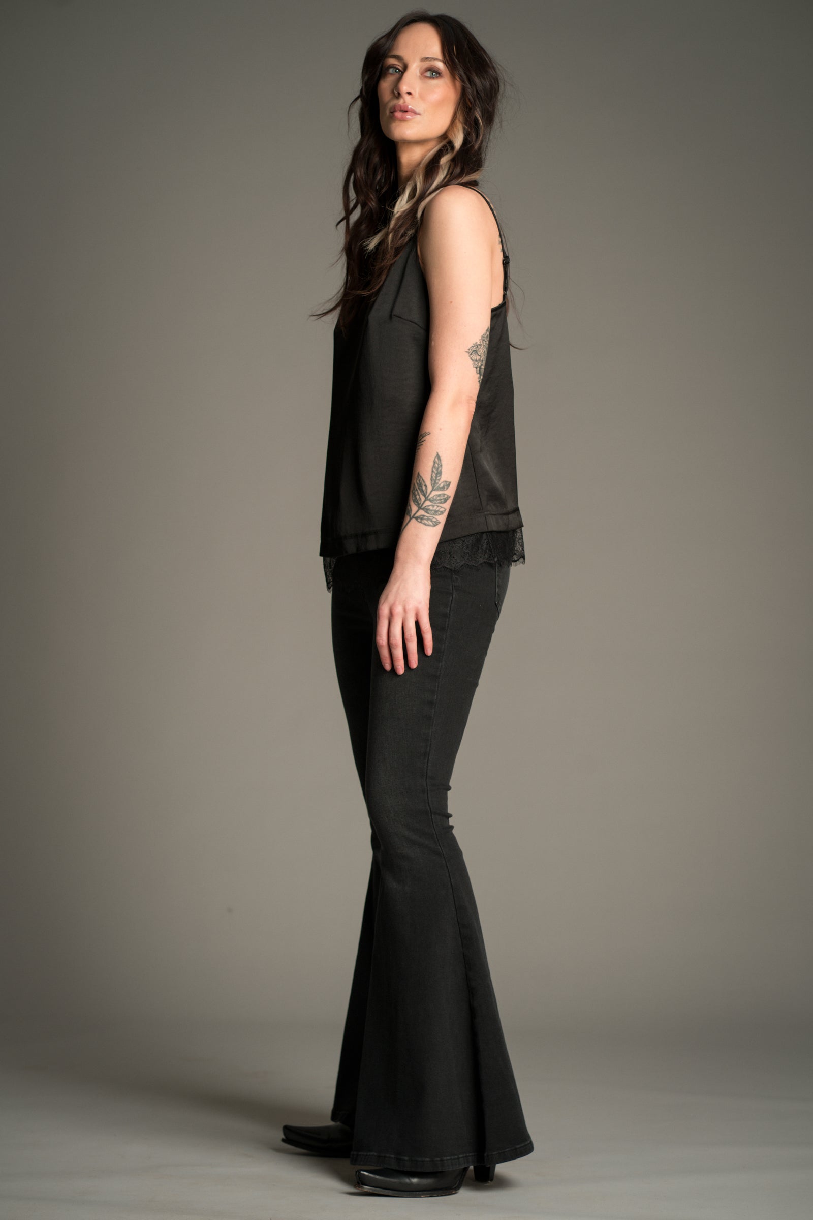 Victoria wears Little Lies Hendrix Denim Flares in washed black denim, side view showing high‑waist fit, subtle fading, and wide flared legs, styled with a black satin cami.