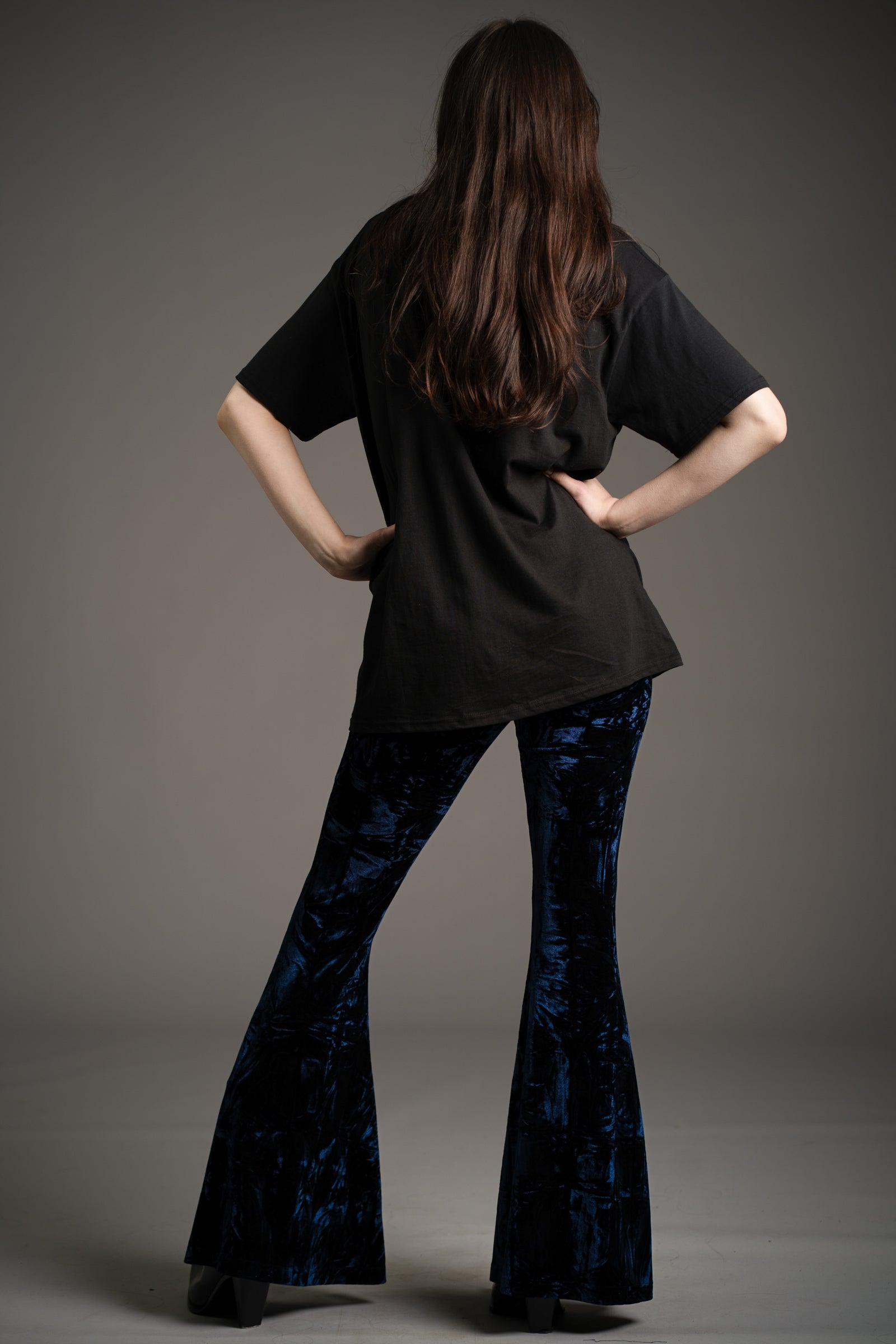 Little Lies Hendrix midnight crushed velvet flared trousers in deep blue, back view showing dramatic bell-bottom silhouette, styled with oversized black graphic tee and platform boots in studio setting