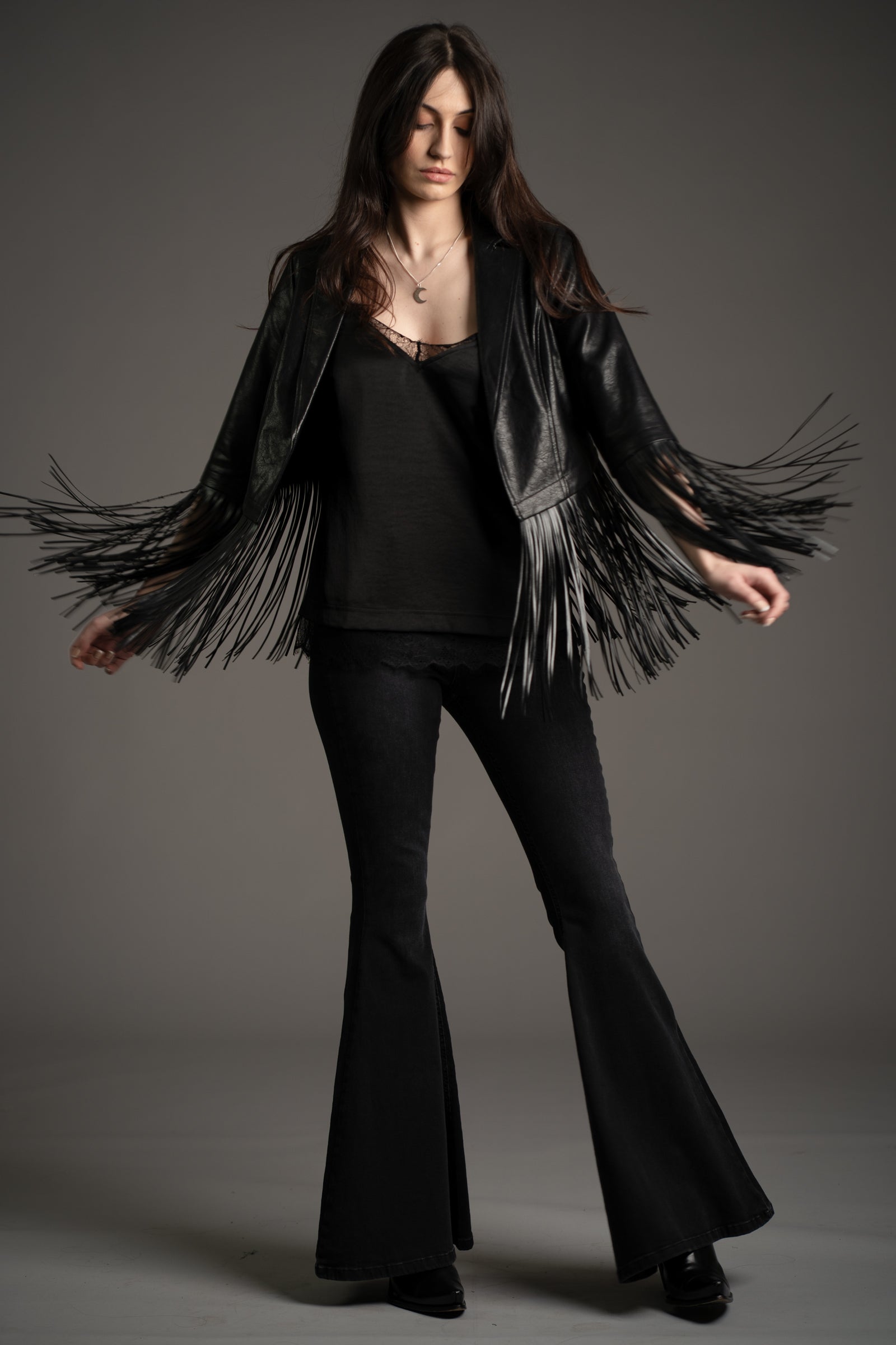 Andie models Little Lies Jagger fringed faux leather jacket in black with dramatic swinging fringe detail, styled with denim flared trousers and cami top