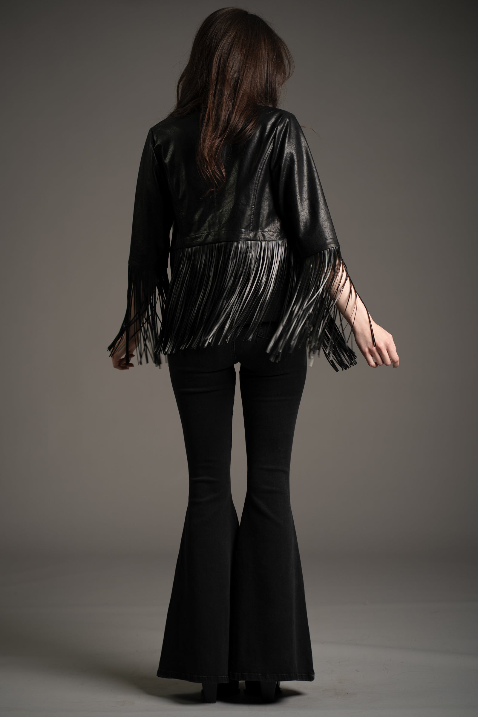 Little Lies Jagger fringed faux leather jacket in black, back view showing long fringe detail, styled with denim flared trousers