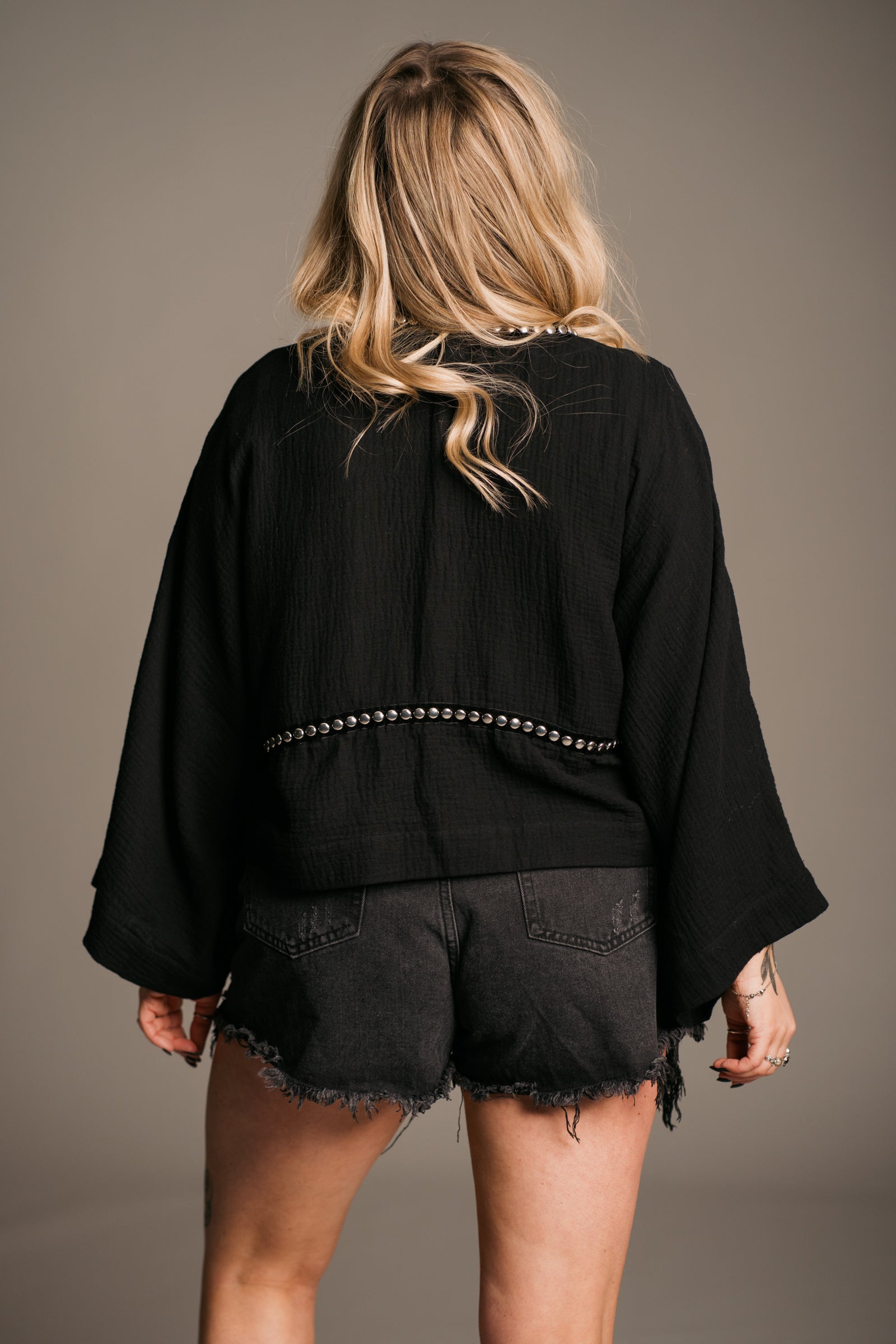 Blonde model Erin wearing the Little Lies Kravitz Black Studded Cheesecloth top showing the back, featuring silver studding along the neckline and front of the Kravitz Studded Top. The top is styled  vintage denim Levi's shorts and paired with a Gypsy Silver Tooth necklace, the look embodies effortless Rock 'n' Roll style