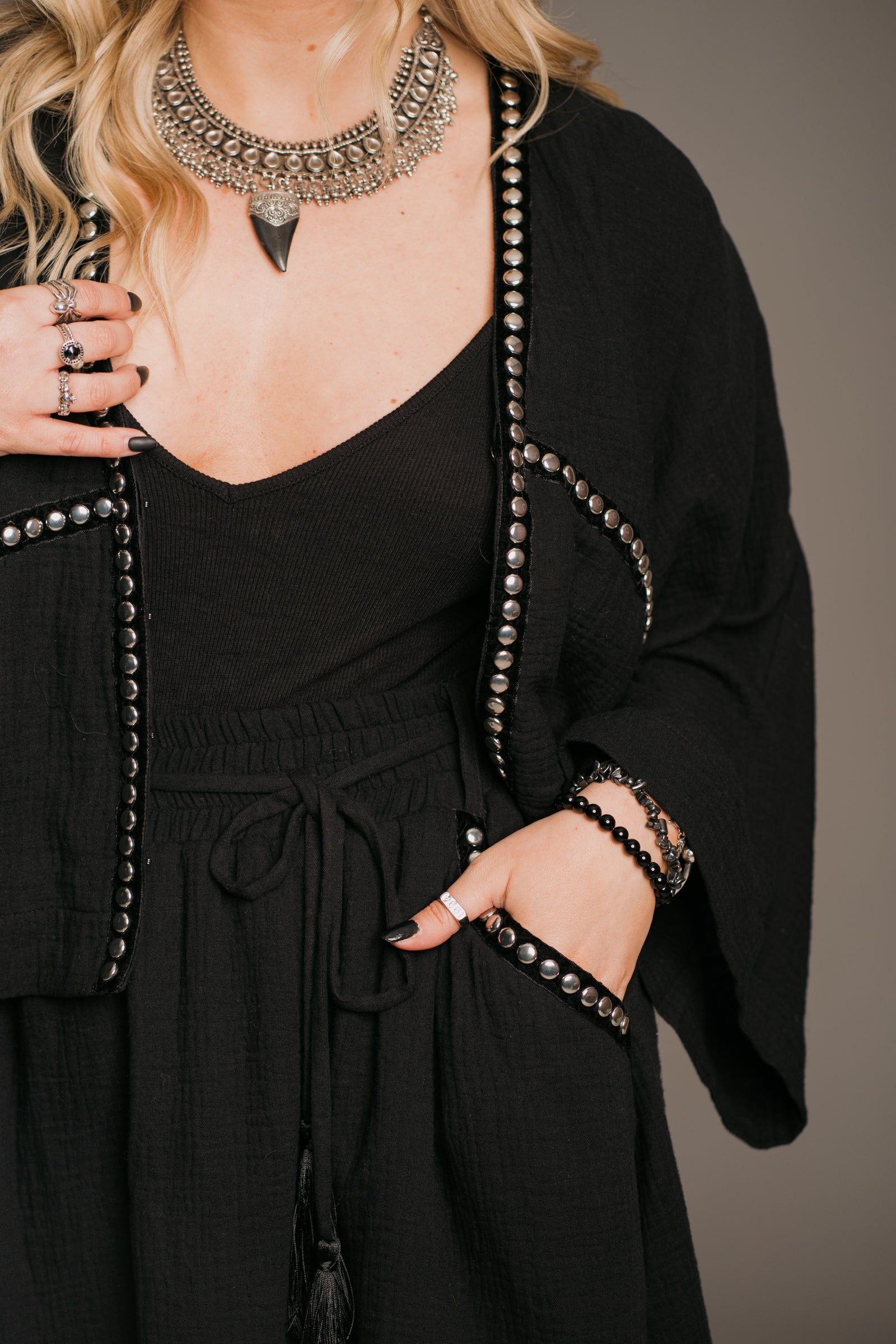 Blonde model Erin wearing the Little Lies Kravitz Black Studded Cheesecloth Co-ord worn open at the front, featuring silver studding along the neckline and front of the Kravitz Studded Top and detailed studding on the hem and pockets of the Kravitz Studded Skort. The skort is styled with a cord tie waist belt and functional pockets. Paired with a Gypsy Silver Tooth necklace, the look embodies effortless Rock 'n' Roll style