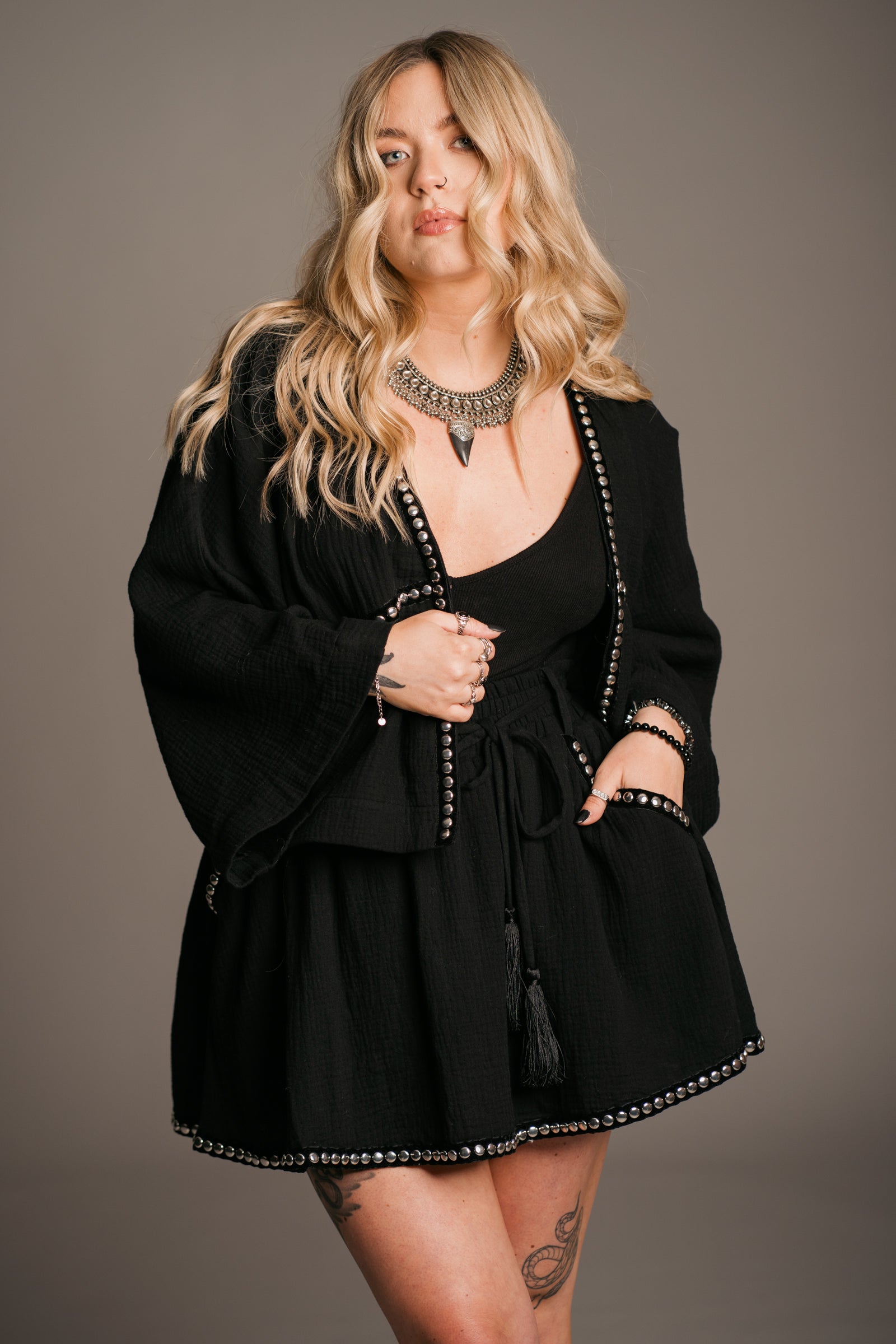 Blonde model Erin wearing the Little Lies Kravitz Black Studded Cheesecloth Co-ord, featuring silver studding along the neckline of the Kravitz Studded Top and detailed studding on the hem and pockets of the Kravitz Studded Skort. The skort is styled with a cord tie waist belt and functional pockets. Paired with a Gypsy Silver Tooth necklace, the look embodies effortless Rock 'n' Roll style