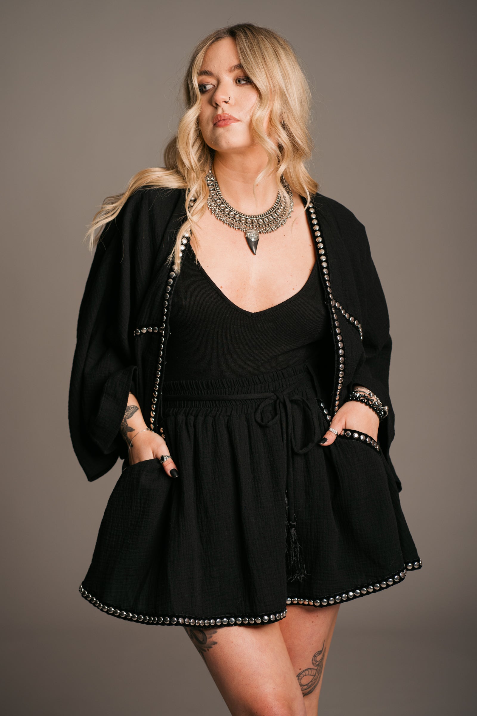 Blonde model Erin wearing the Little Lies Kravitz Black Studded Cheesecloth Co-ord with the top worn open, featuring silver studding along the neckline of the Kravitz Studded Top and detailed studding on the hem and pockets of the Kravitz Studded Skort. The skort is styled with a cord tie waist belt and functional pockets. Paired with a Gypsy Silver Tooth necklace, the look embodies effortless Rock 'n' Roll style