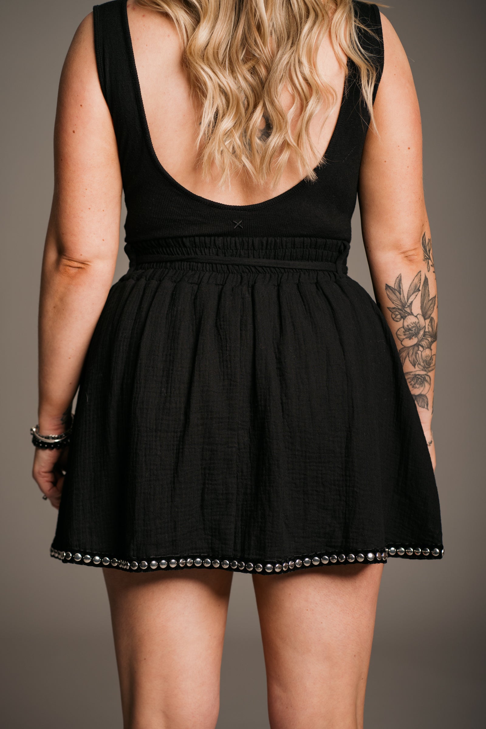 Blonde model Erin wearing the Little Lies Kravitz Black Studded Cheesecloth Skort showing the back, featuring silver stud details on the hem and pockets of the Kravitz Studded Skort. The skort has an elasticated chirred waist with a cord tie belt and functional pockets. Paired with a simple black vest top and a Gypsy Silver Tooth necklace, the look embodies effortless Rock 'n' Roll style
