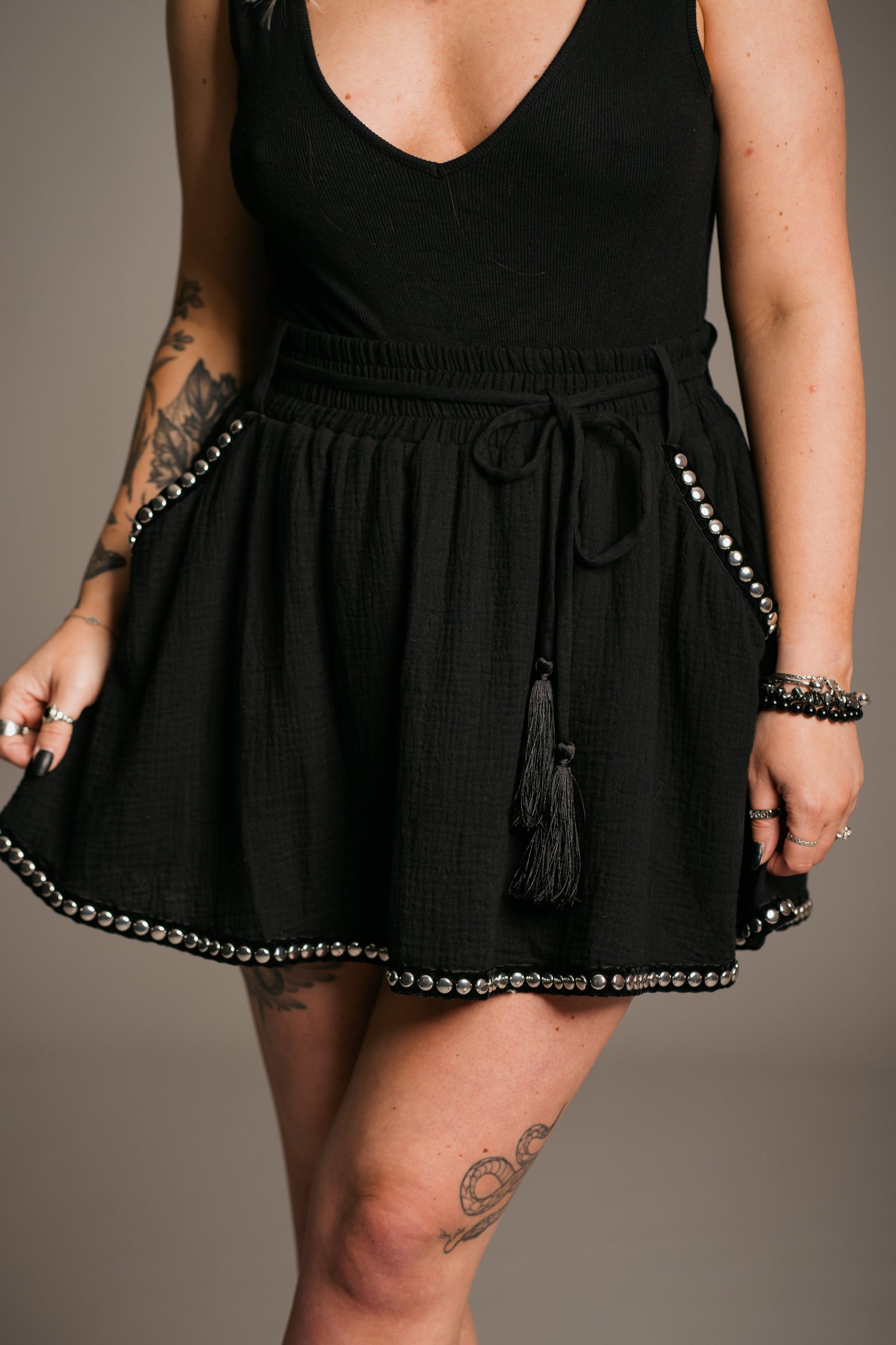Blonde model Erin wearing the Little Lies Kravitz Black Studded Cheesecloth Skort, featuring silver stud details on the hem and pockets of the Kravitz Studded Skort. The skort has an elasticated chirred waist with a cord tie belt and functional pockets. Paired with a simple black vest top and a Gypsy Silver Tooth necklace, the look embodies effortless Rock 'n' Roll style