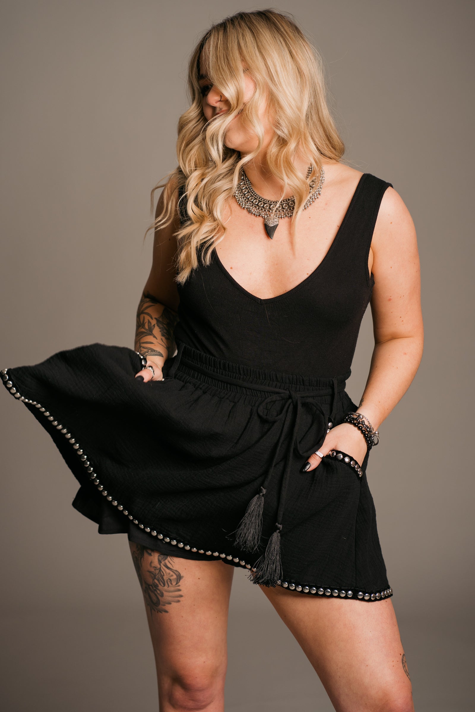 Blonde model Erin wearing the Little Lies Kravitz Black Studded Cheesecloth Skort, featuring silver stud details on the hem and pockets of the Kravitz Studded Skort. The skort has an elasticated chirred waist with a cord tie belt and functional pockets. Paired with a simple black vest top and a Gypsy Silver Tooth necklace, the look embodies effortless Rock 'n' Roll style