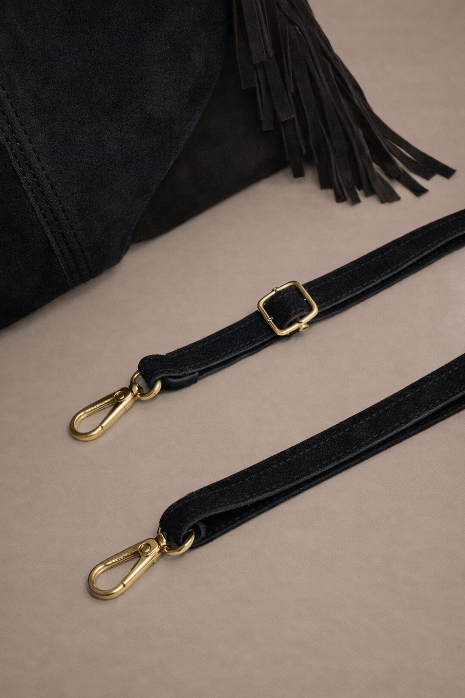 Little Lies Lila black genuine suede bag with detachable strap showing gold hardware and buckle detail in studio setting.