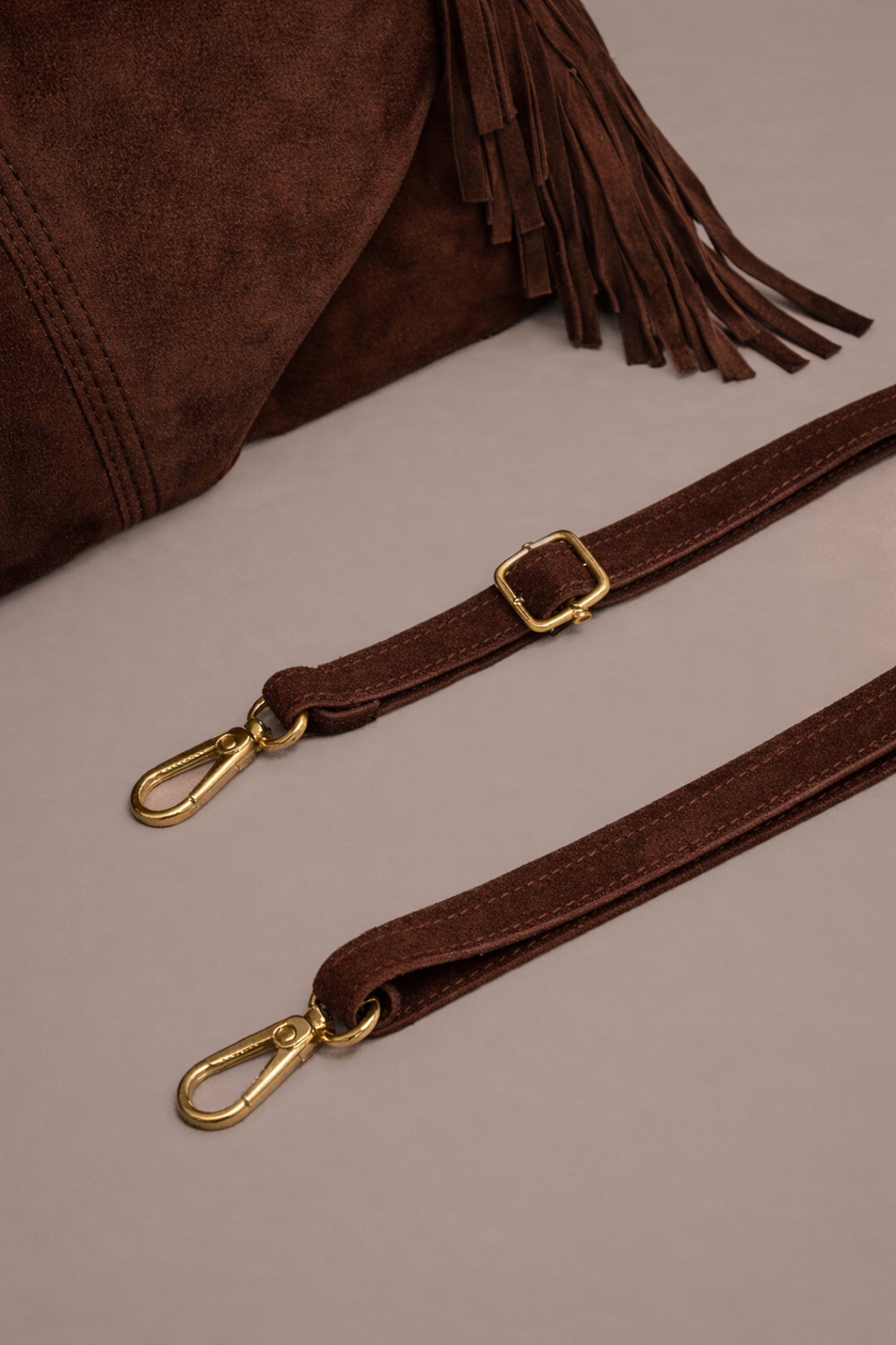 Little Lies Lila chocolate genuine suede bag with detachable strap showing gold hardware and buckle detail in studio setting.