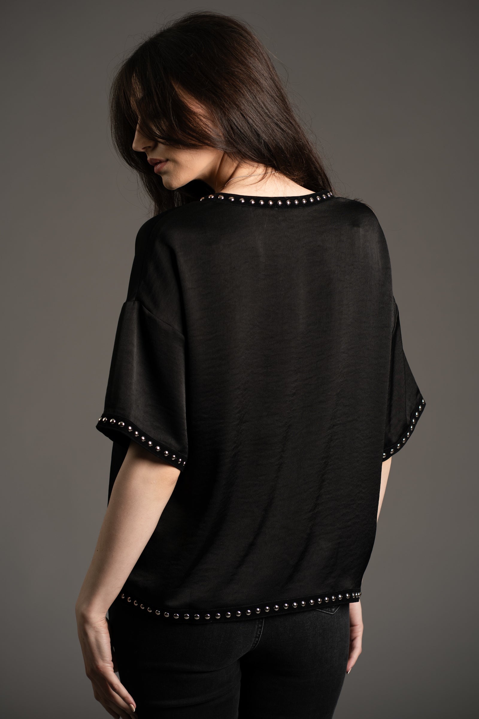 Little Lies Lita black satin tee with silver studded trim on neckline, sleeves, and hem shown from back in studio setting