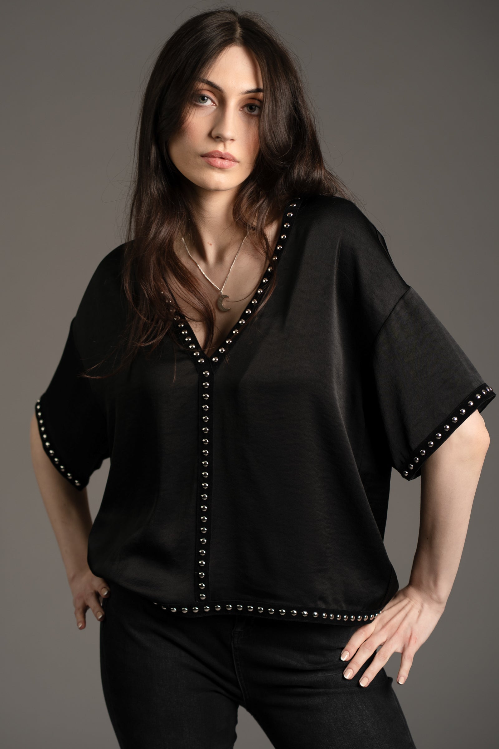 Andie models Little Lies Lita black satin tee with silver studded trim on neckline, sleeves, and hem styled with black skinny jeans in studio setting