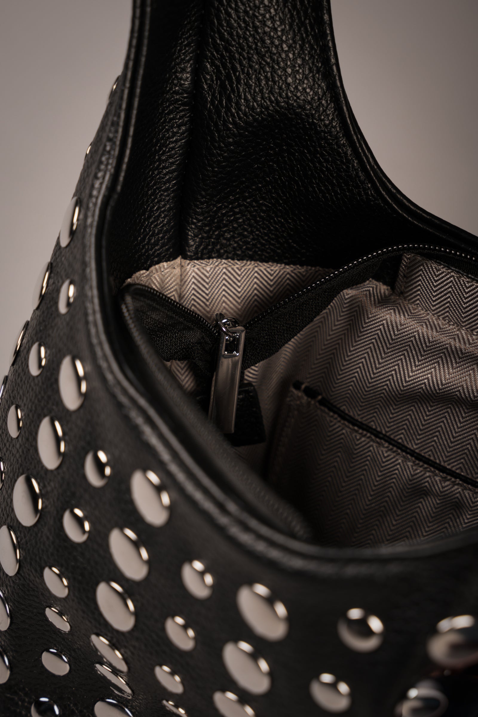 Close-up of Little Lies Viva black studded leather handbag showing zip closure and interior lining with herringbone pattern, studio setting.