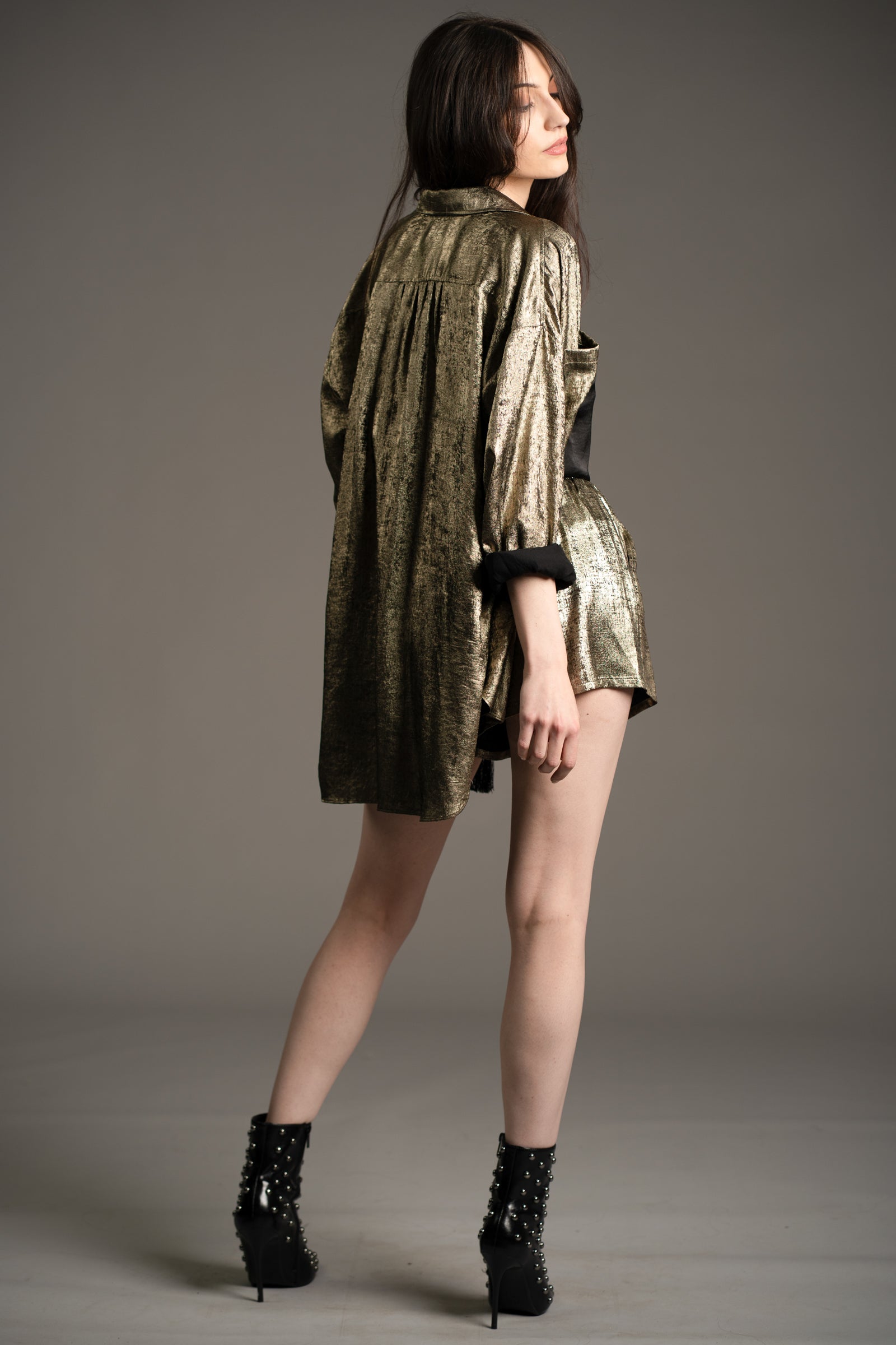 Andie models Little Lies Luna metallic shirt in gold styled for party season with matching metallic shorts and studded ankle boots, back view in studio setting