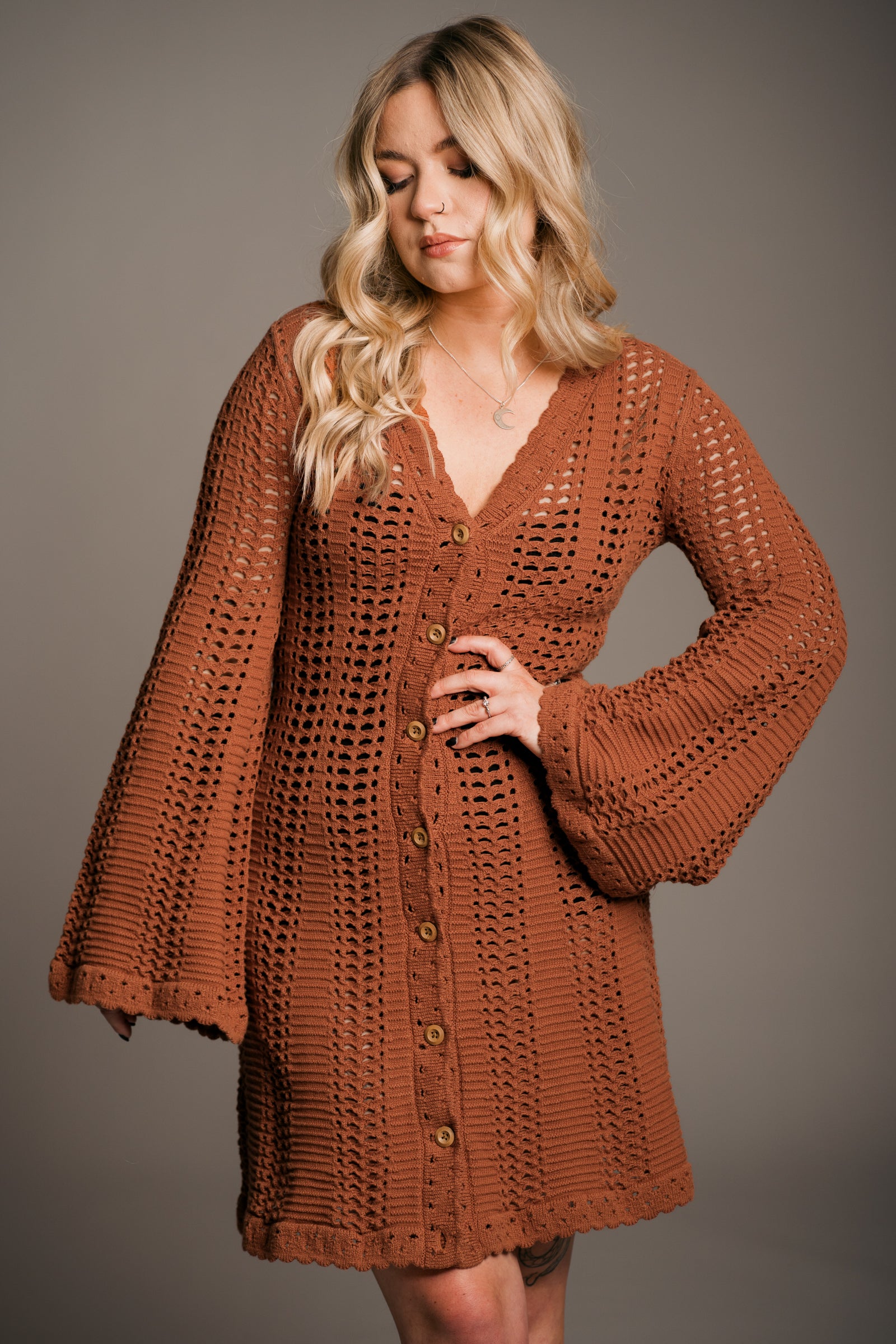 Our model, Erin, stands against a neutral studio backdrop, wearing the Little Lies Marianne Rust Crochet Knit Mini Dress. The dress features intricate crochet detailing, a full button front, a V-neckline, and flared sleeves—exuding a relaxed bohemian aesthetic.