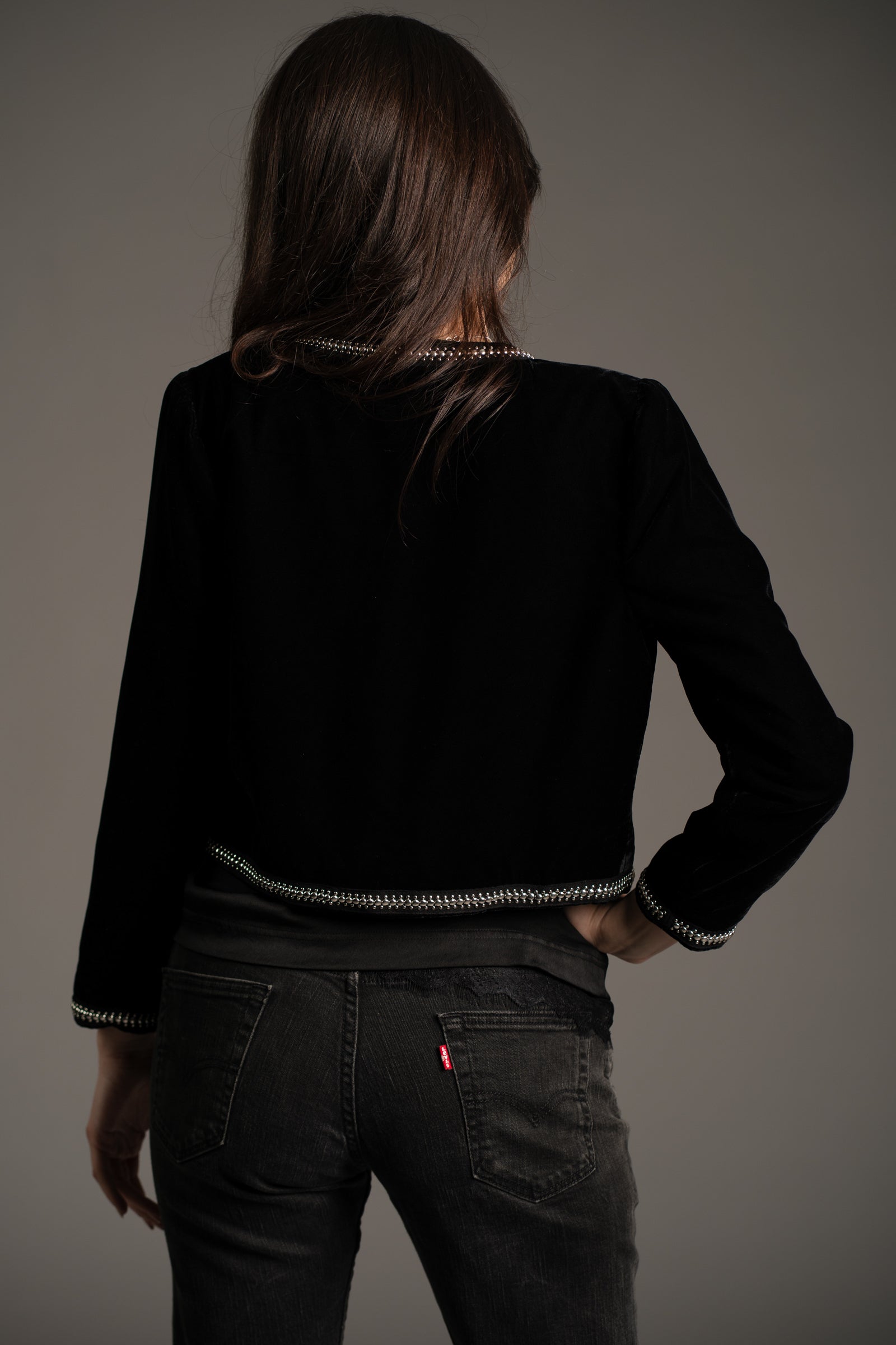 Little Lies Metal Guru studded velvet jacket in black, back view showing silver stud detailing on hem and cuffs, styled with dark denim jeans in studio setting