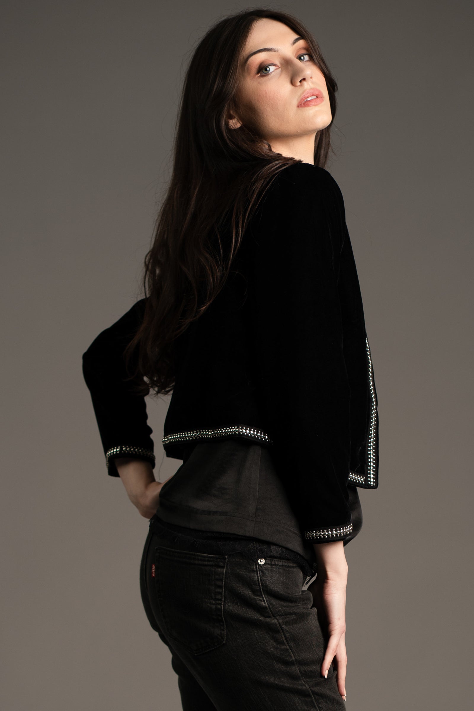 Andie models Little Lies Metal Guru studded velvet jacket in black, side-back view showing silver stud trim on hem and cuffs, styled with dark jeans in studio setting
