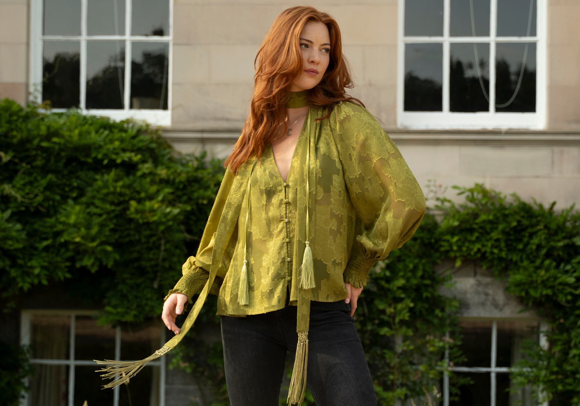 Anna wearing the Little Lies Cassia Olive Chiffon Smock Top with long tassel ties, styled with black denim, standing outdoors in front of a historic stone building covered in greenery.