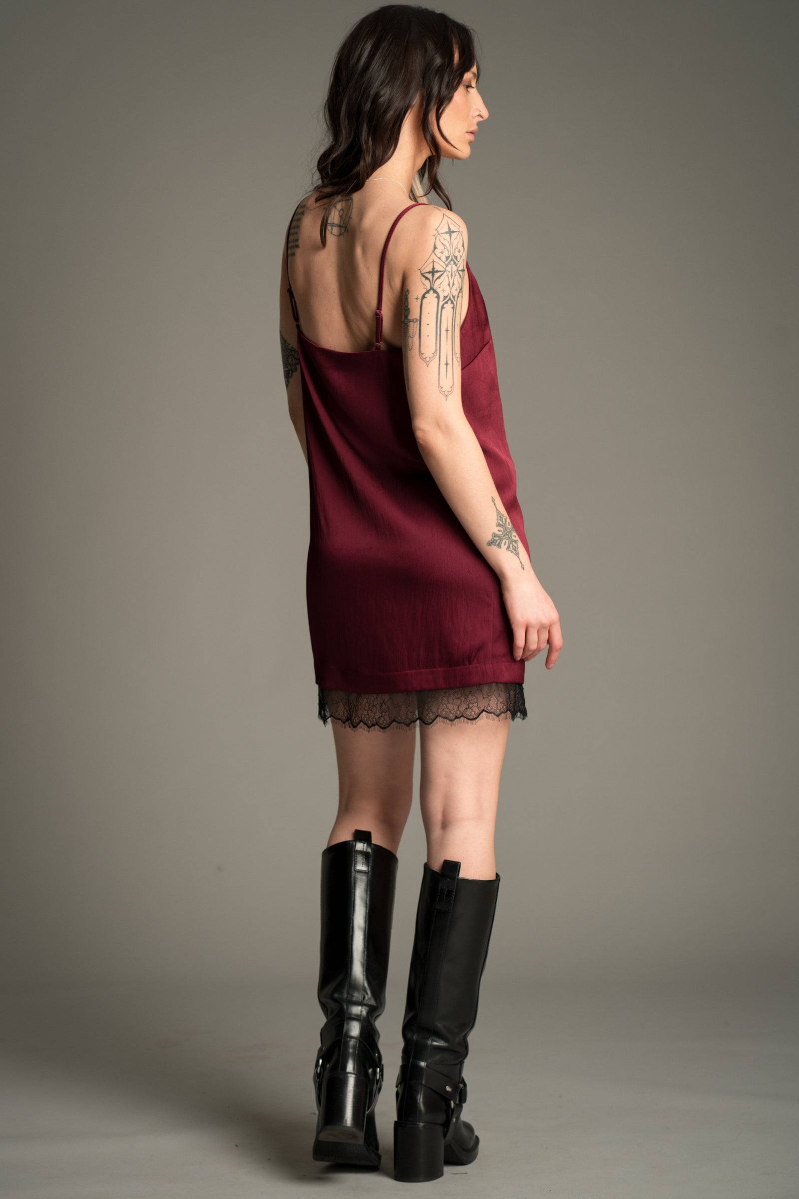 Victoria posing against a grey studio backdrop, wearing the Little Lies Nightbird Plum Satin Slip Dress Mini showing the back of the dress with lace trim hem