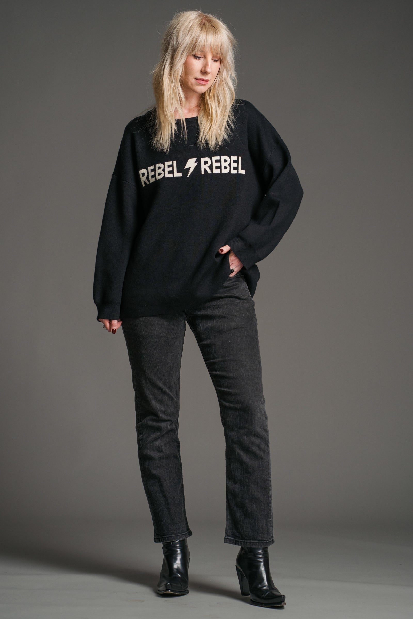 Sara wearing a the Rebel Rebel Black Graphic Jumper with 'REBEL REBEL' ivory text and lightning bolt graphic on a gray background