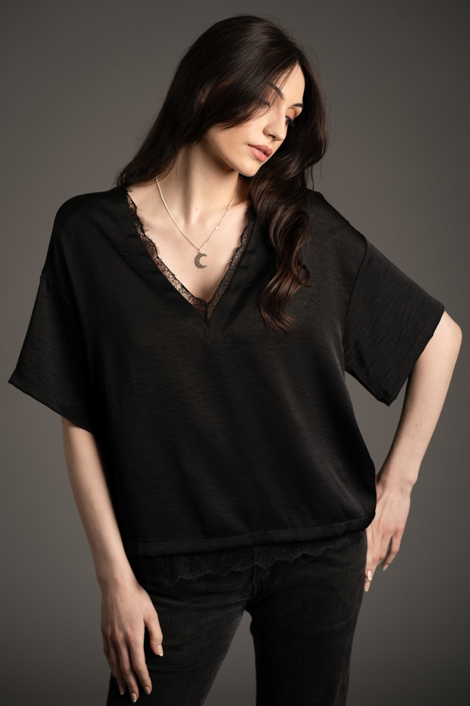 Andie models Little Lies Roxy lace trim satin tee in black with deep V-neckline and delicate lace edging, styled with black jeans in studio setting