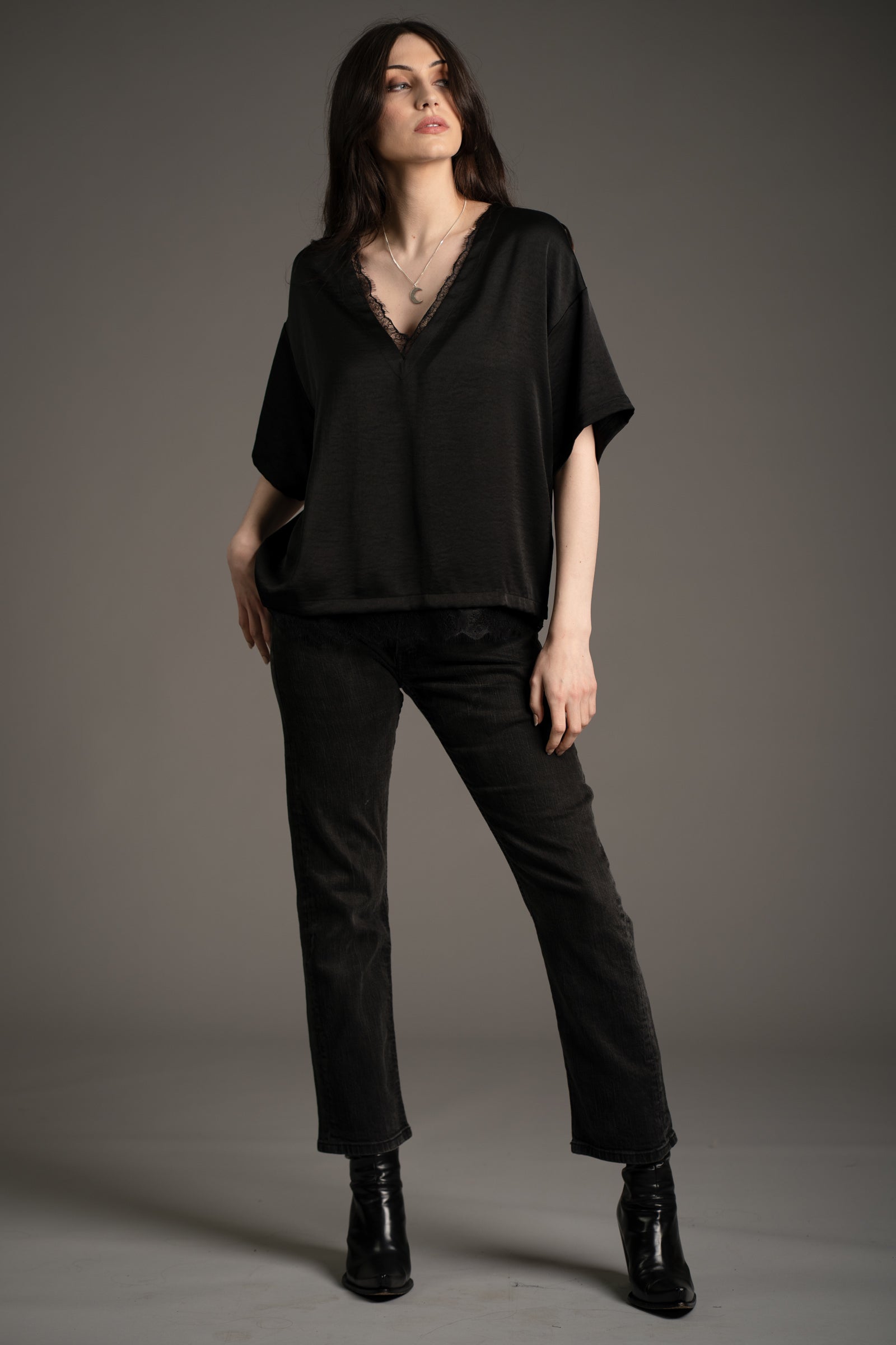 Andie models Little Lies Roxy lace trim satin tee in black with deep V-neckline and delicate lace edging, styled with straight-leg black jeans and black ankle boots in studio setting