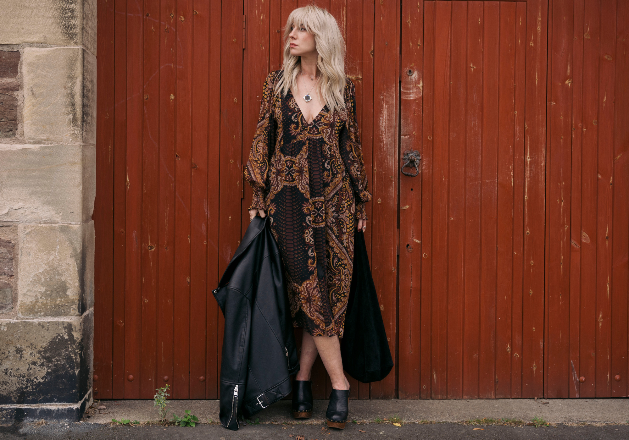 Sara stood in front of a brown door wearing Little Lies Peacekeeper Midi Dress, holding a Olsen Black Suede slouchy bag and black leather jacket.