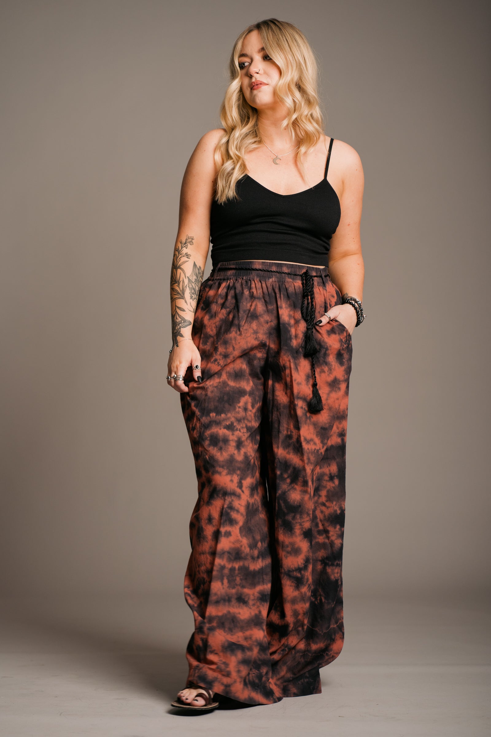 Blonde model Erin wearing the Little Lies Zephyr Tie Dye Co-Ord trousers, a vibrant tie-dye wide leg design with a black tie waist cord and pockets. Styled with the Little Lies sterling silver crescent moon necklace a simple black vest top and ankle-tie black sandals, the look embodies effortless bohemian style against a neutral backdrop.