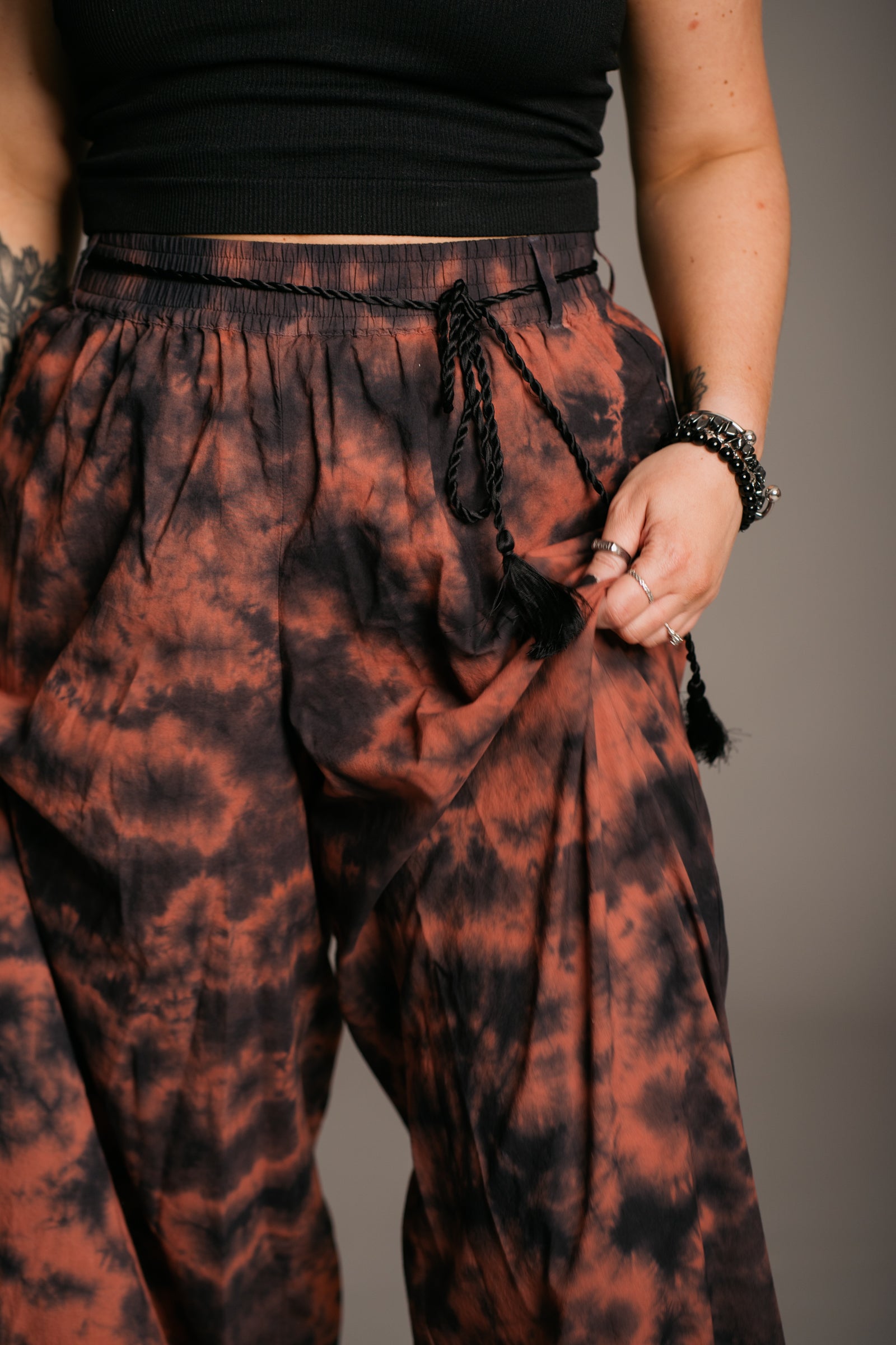 Model Erin wearing the Little Lies Zephyr Tie Dye Co-Ord trousers, a vibrant tie-dye wide leg design with a black tie waist cord and pockets. Styled with a simple black vest top and ankle-tie black sandals, the look embodies effortless bohemian style against a neutral backdrop.