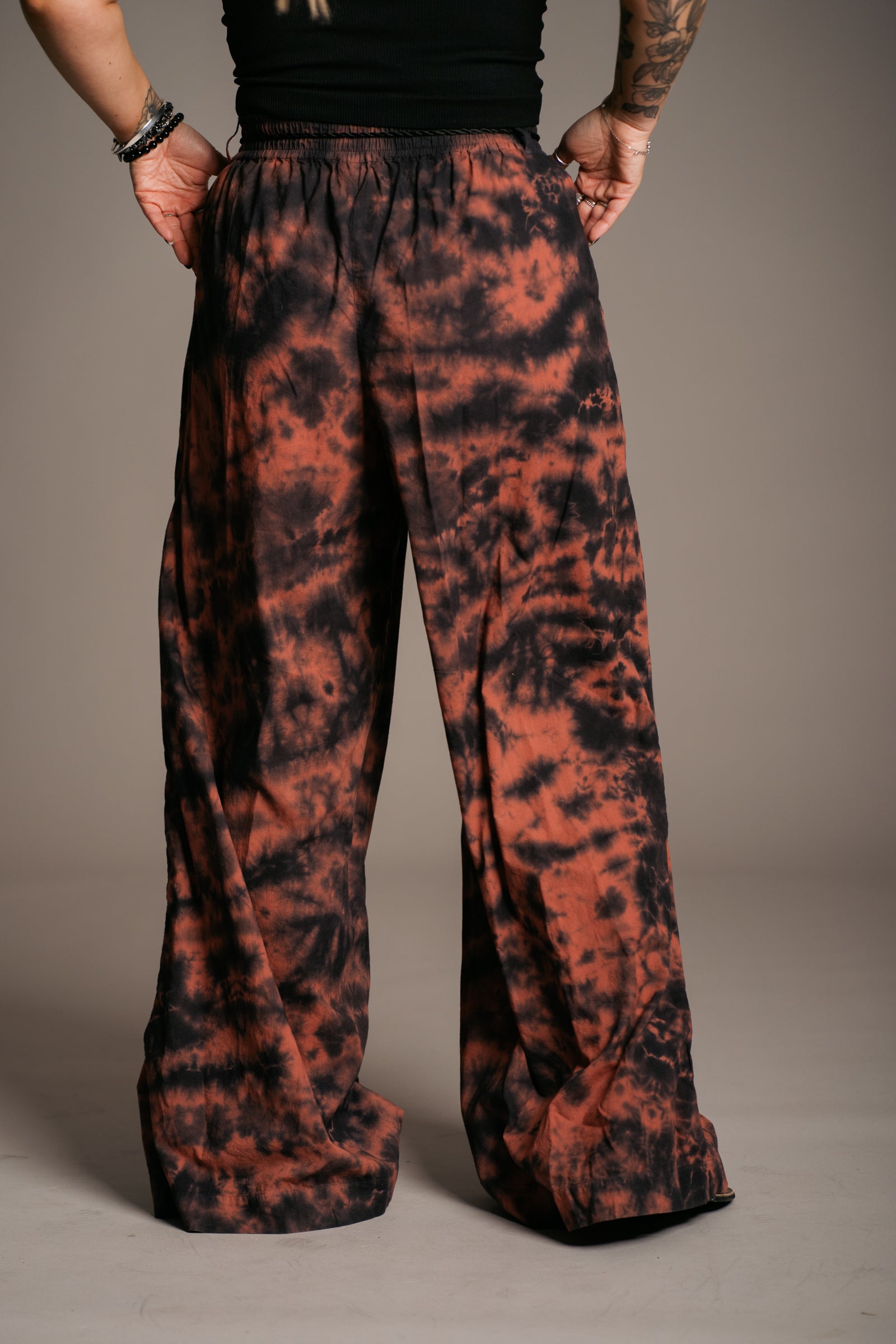 Model Erin wearing and showing the back of the Little Lies Zephyr Tie Dye Co-Ord trousers, a vibrant tie-dye wide leg design with a black tie waist cord. Styled with a simple black vest top and ankle-tie black sandals, the look embodies effortless bohemian style against a neutral backdrop.