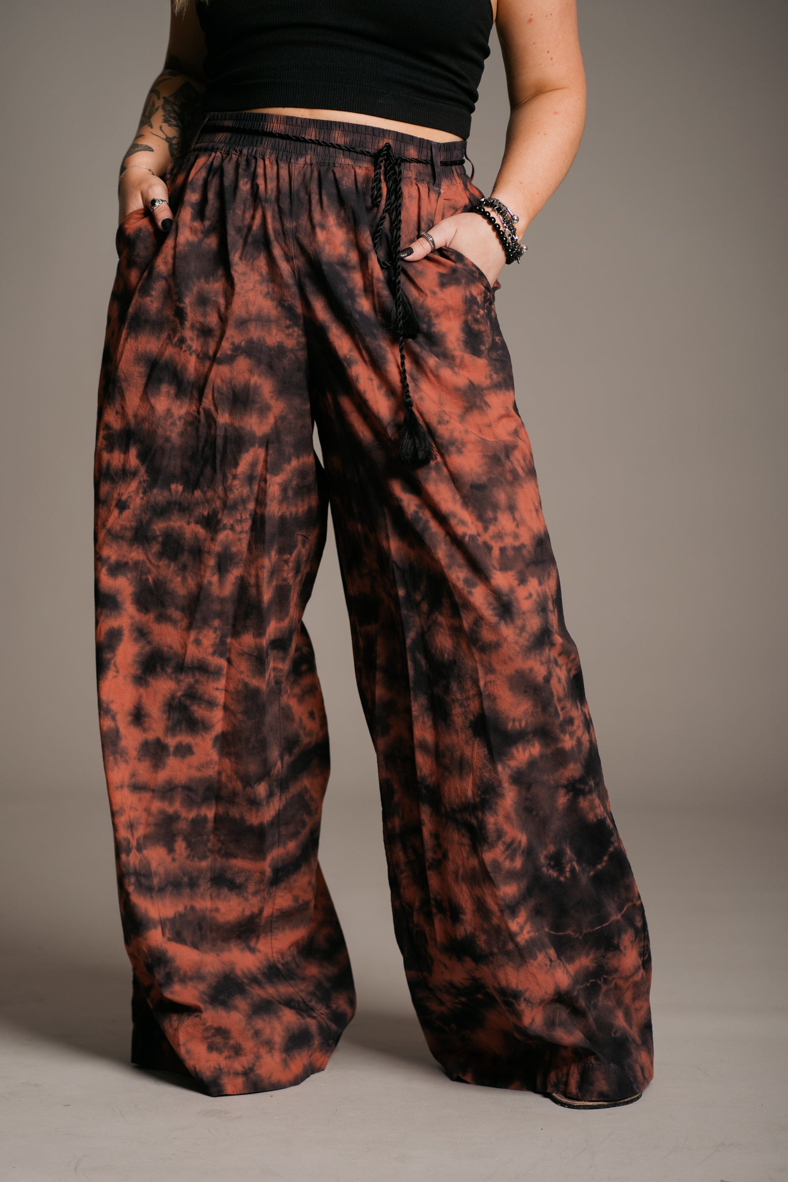 Model Erin wearing the Little Lies Zephyr Tie Dye Co-Ord trousers, a vibrant tie-dye wide leg design with a black tie waist cord and pockets. Styled with a simple black vest top and ankle-tie black sandals, the look embodies effortless bohemian style against a neutral backdrop.