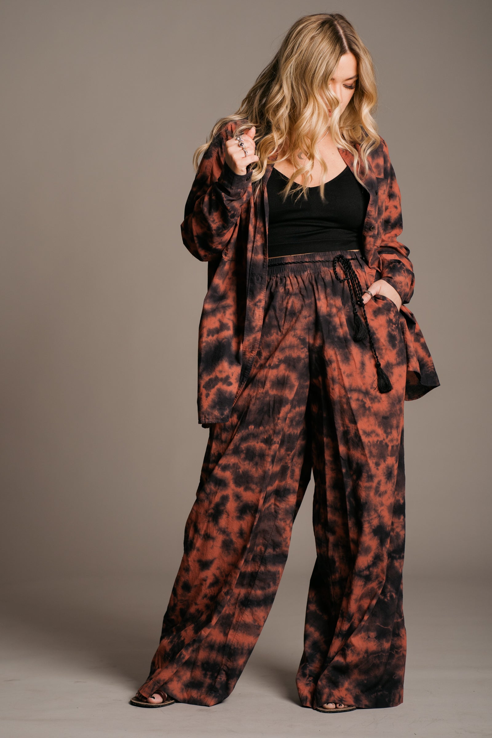 Model Erin wearing the Little Lies Zephyr Tie Dye Co-Ord, a vibrant tie-dye co-ord set featuring a relaxed-fit long-sleeve shirt and matching wide-leg, tie-waist trousers. Styled with ankle-tie black sandals, the look embodies effortless bohemian style against a neutral backdrop.