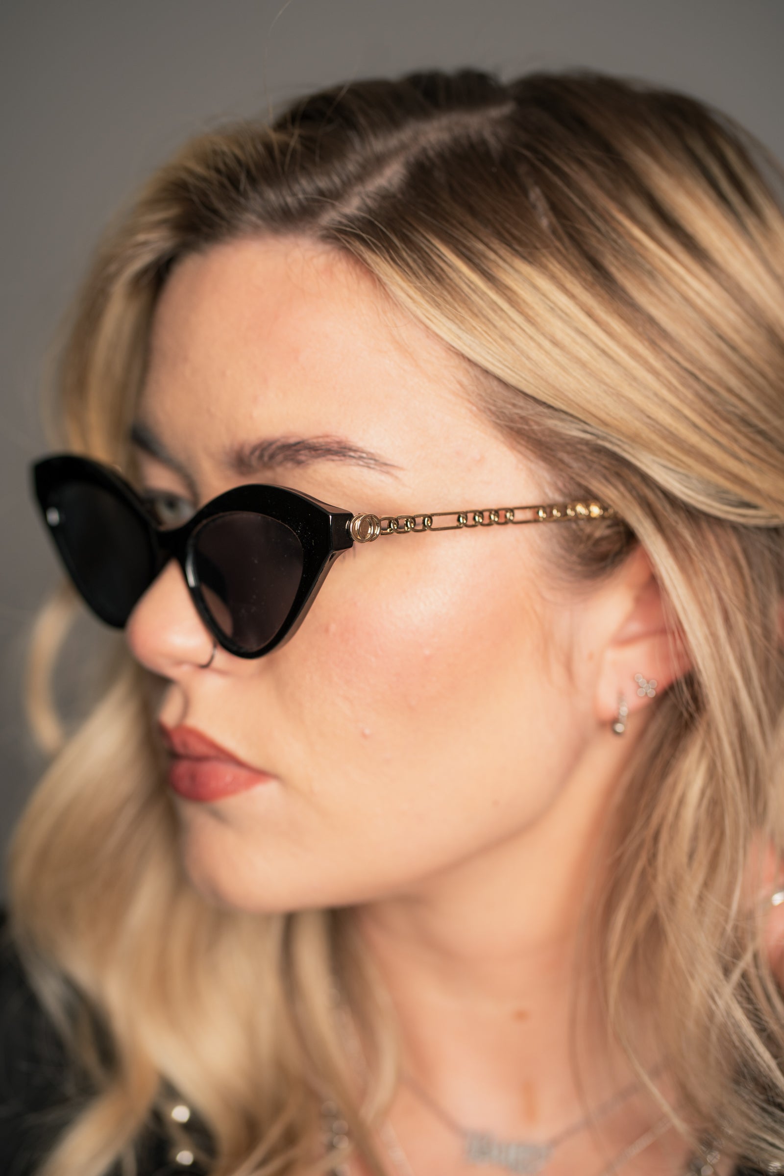 Little Lies Zoe black cat‑eye sunglasses with gold chain arm detail and dark tinted lenses, retro frame, side close‑up view