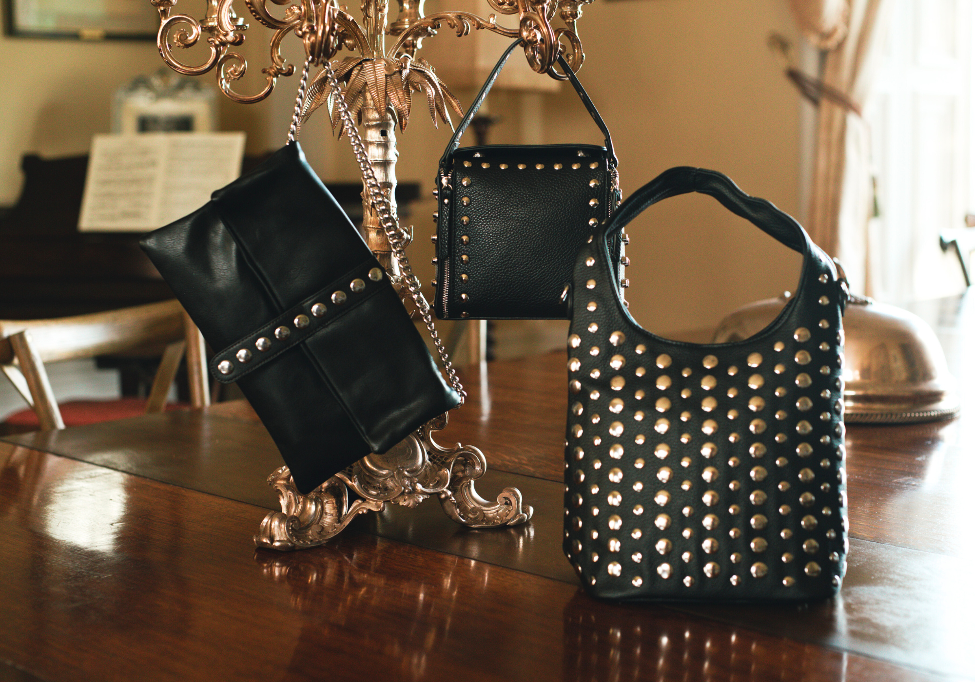 Little Lies Viva black studded hobo bag, Leo black studded box bag, and Cleo chain clutch bag displayed on ornate gold stand in a vintage-inspired setting