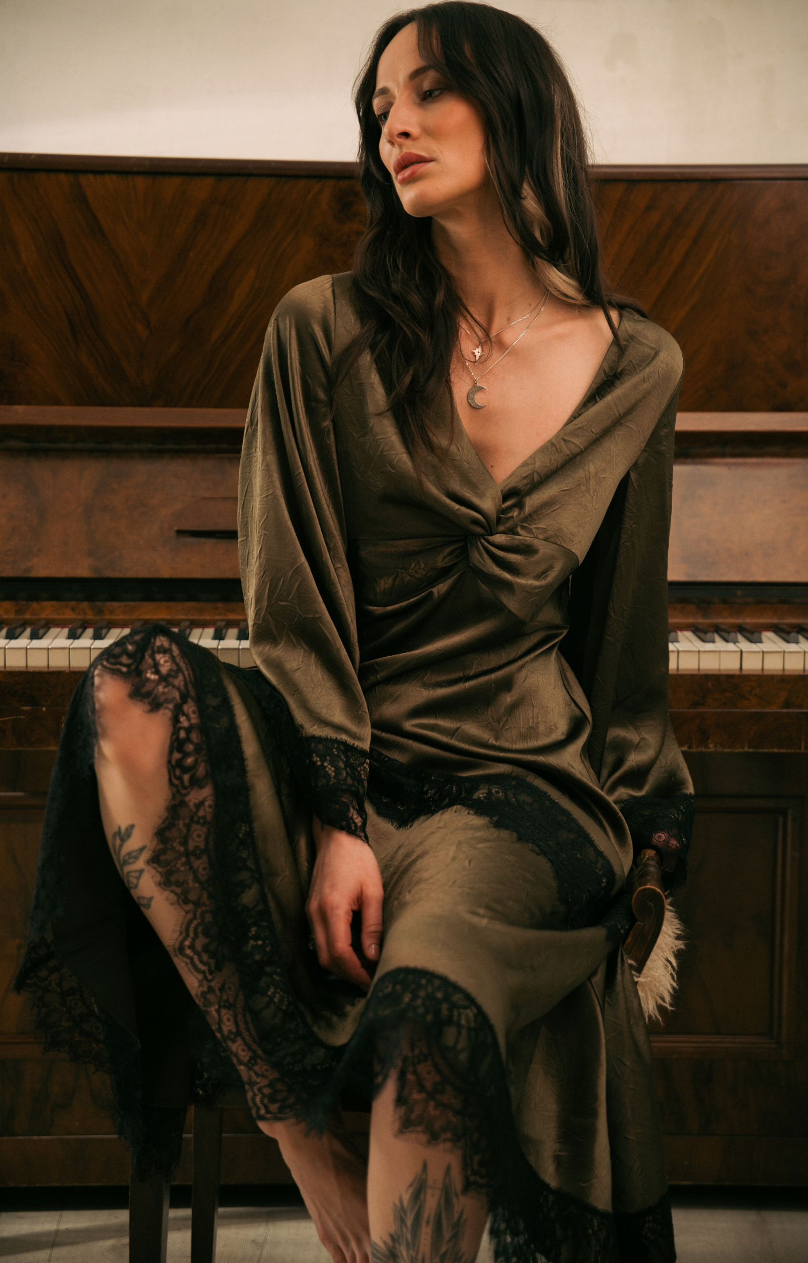 Full‑length view of Model in the Little Lies Arabella Satin Maxi Dress in olive satin with black lace trim, featuring flared sleeves and an asymmetric hem.