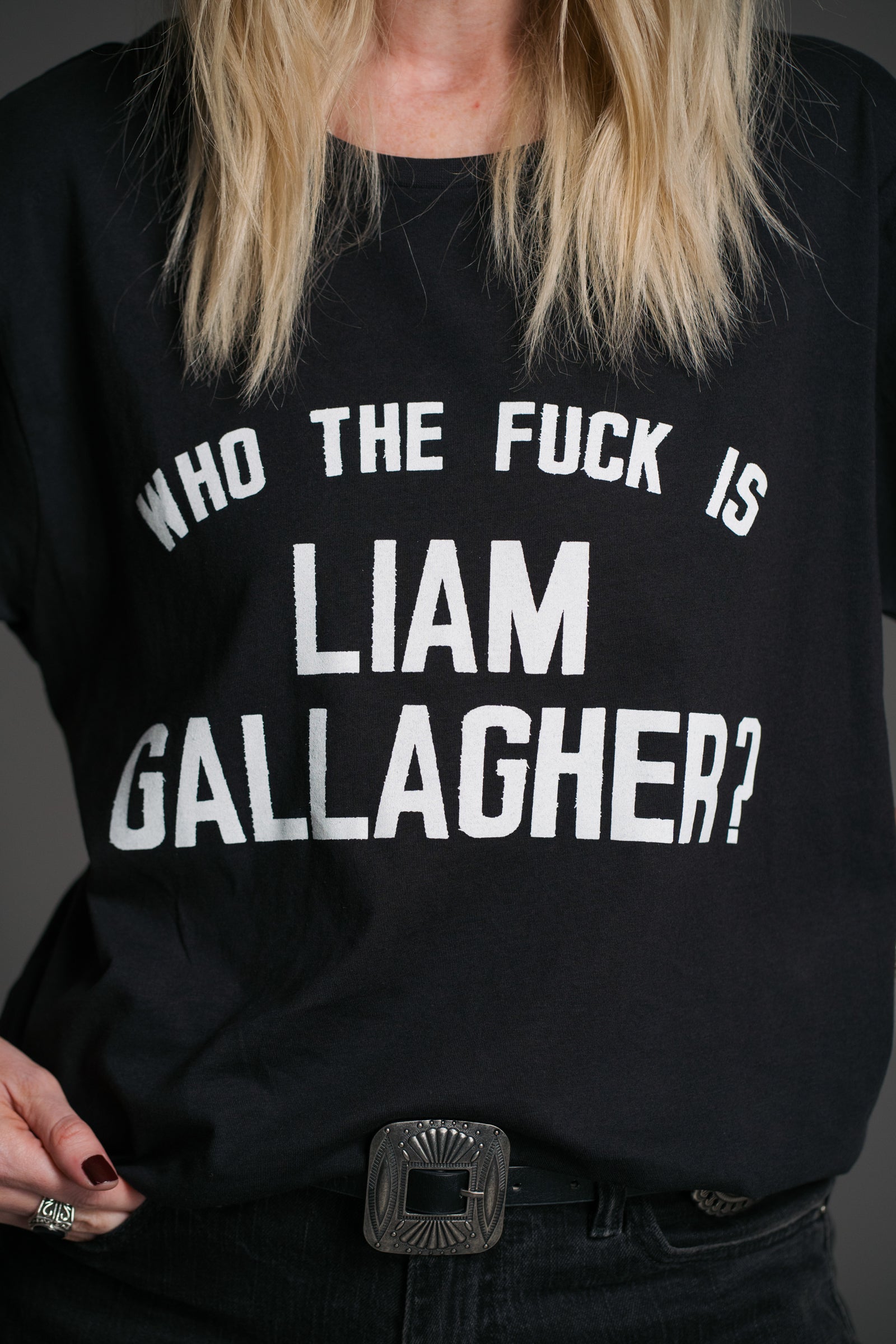 Unisex black Liam Gallagher graphic slogan t‑shirt by Little Lies, close‑up view with western‑style belt and silver jewellery
