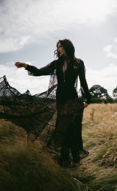 Model wearing the Little Lies Rhiannon Black Maxi Dress in a Spellbound collection campaign shoot, featuring sheer lace detailing and flowing black silhouette in a countryside setting.