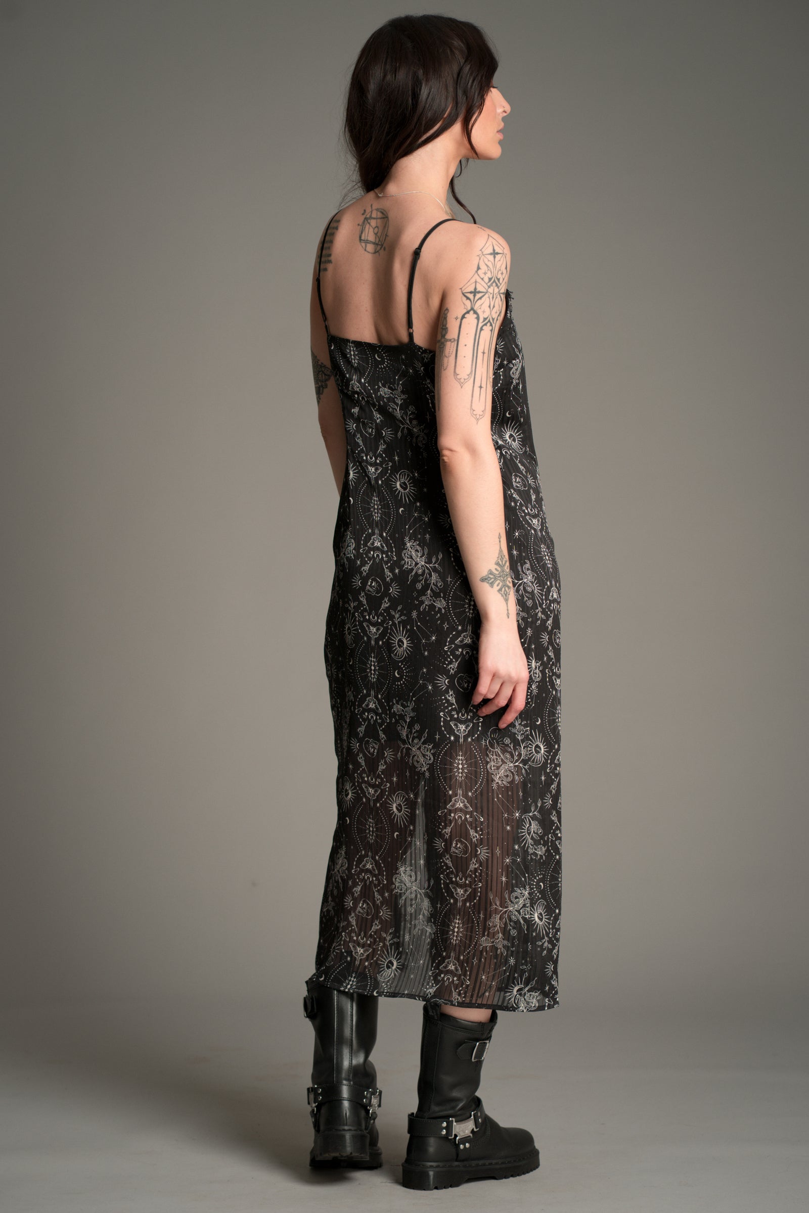 Side‑back view of the Little Lies Tattoo Slip Dress in black patterned lace with sheer detailing and adjustable straps, styled with chunky black boots.