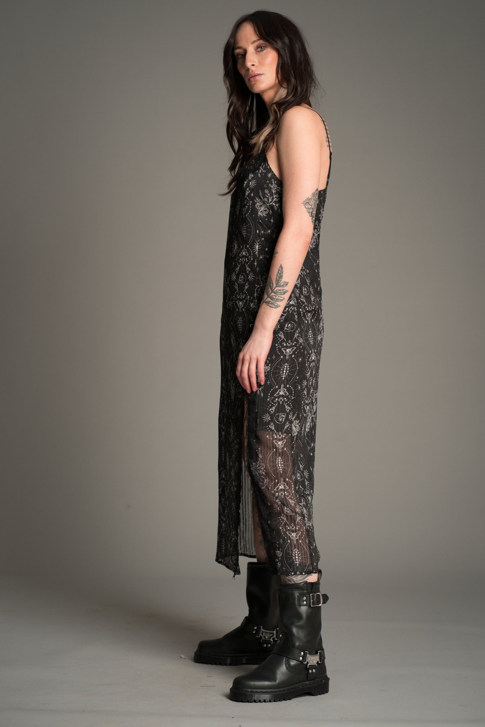 Side view of Victoria in the Little Lies Tattoo Slip Dress in black patterned lace with sheer detailing and a side split, styled with chunky black buckle boots.